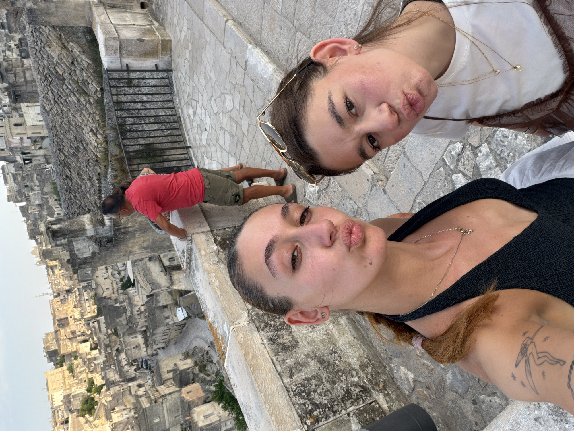 In Matera, Italy, two young women are posing for a selfie with a pout. The woman on the left has a black halter top and a tattoo on her arm. The woman on the right is wearing a white t-shirt and has sunglasses perched on her head. In the background, a man in a red shirt and khaki shorts stands near a railing overlooking the ancient city of Matera, a UNESCO World Heritage site known for its cave dwellings. The city's architecture, with its stone buildings cascading down the hillside, is visible behind them.