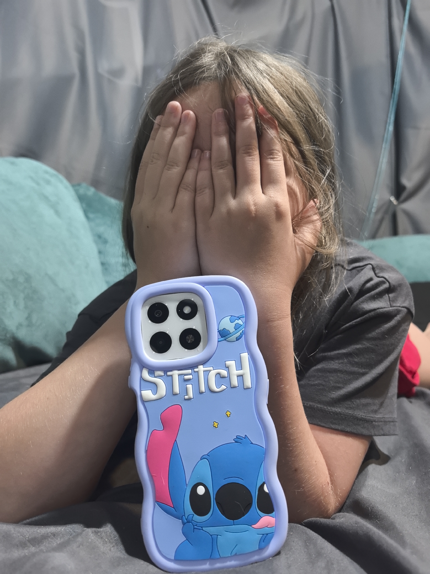 The image shows a close-up of a young person covering their face with their hands. The person is wearing a grey t-shirt and has light brown hair. In front of them is a light purple phone case decorated with the character Stitch from Disney's Lilo & Stitch. The phone case is held by the person's arm, which is visible in the lower left portion of the image. The background consists of a grey textured surface, likely a bed or sofa, and a teal-colored cushion. There is also a hint of a grey curtain in the upper background. There is no discernible information about the location within Noviy, Russia, as the image lacks any identifying landmarks or context.