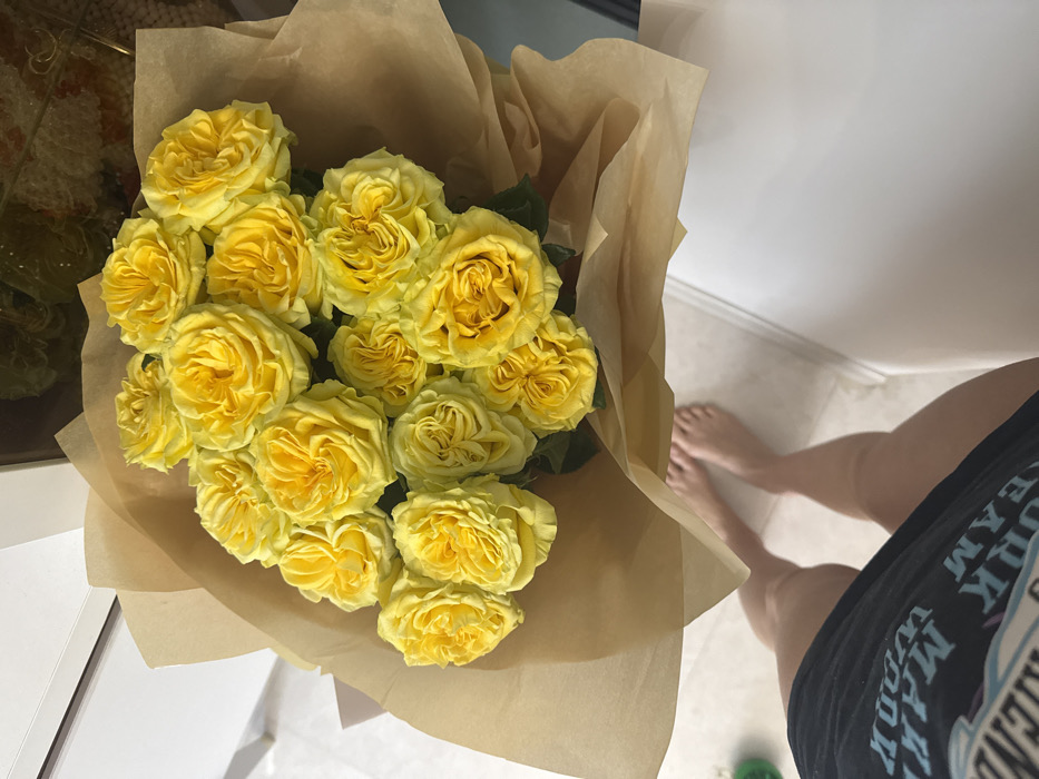 The image displays a bouquet of approximately 15-20 bright yellow roses, wrapped in plain brown kraft paper, held or positioned at chest height relative to the viewer. The roses are in full bloom, exhibiting a vibrant yellow hue. In the lower portion of the frame, the bare feet and lower legs of an individual are visible, standing on a light-colored tiled floor. A dark garment with light-colored, inverted text is partially visible on the individual's upper left leg. The background consists of light-colored interior surfaces, including a white horizontal counter or table on the right and a white vertical surface on the left. A blurred, translucent container holding light-colored decorative elements is present in the upper right background. The scene is indoors, and no specific landmarks or external features are present to identify a precise location within Batumi, Georgia; it appears to be a residential or commercial interior.