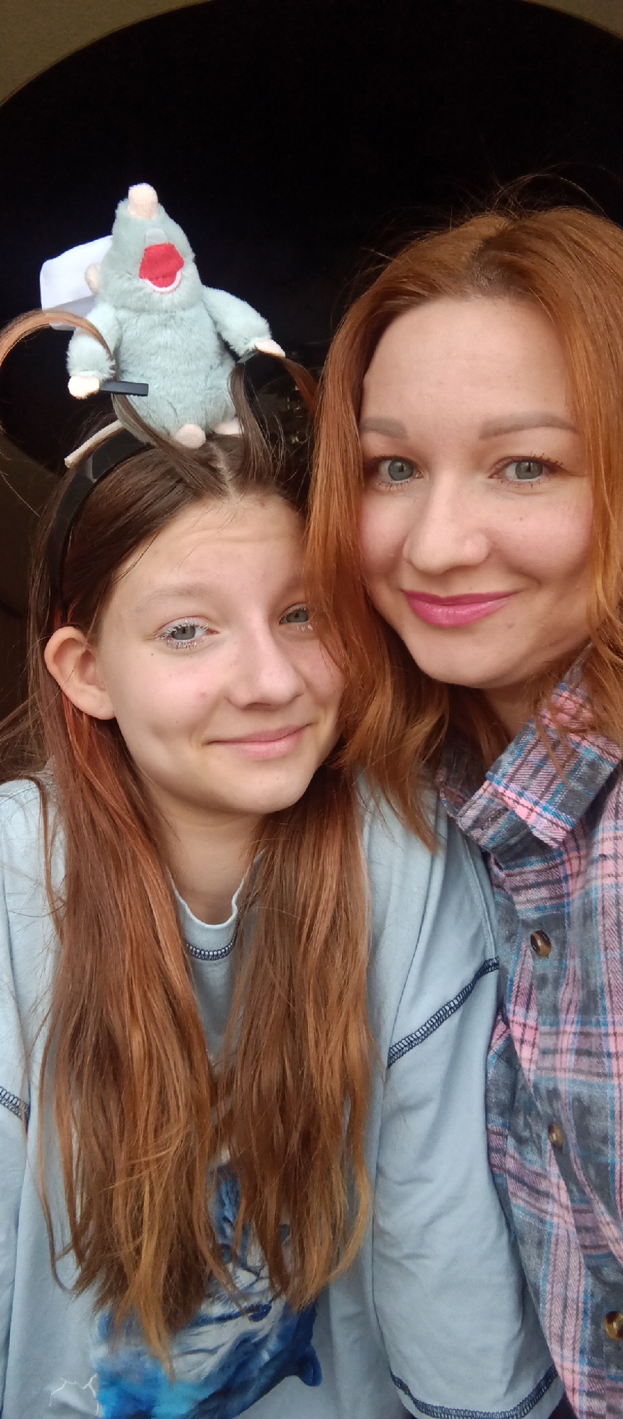 A close-up selfie shows two females, likely a mother and daughter, smiling at the camera. The woman on the right has reddish-orange hair and bright blue-green eyes, wearing light pink lipstick and a plaid shirt with pink, grey, and blue patterns. Her smile is broad and friendly.
The younger female on the left has long, reddish-brown hair and light blue eyes, with what appears to be white or silver glitter on her eyelashes. She is wearing a light blue long-sleeved shirt with a partial print of a blue cat visible towards the bottom. On top of her head, attached to a black headband, is a small, light green plush toy resembling a mole or mouse, featuring a prominent red mouth and small white or light pink ears and nose.
Both subjects have pleasant, smiling expressions. The background is dark and indistinct, suggesting an indoor setting or a car interior. No specific location cues or visible text are present. The overall mood is cheerful and intimate.