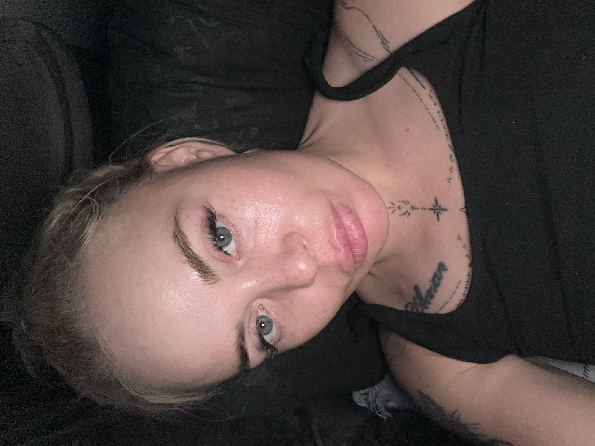 A medium shot, eye-level, selfie features a Caucasian woman with blonde hair pulled back in a bun. She is wearing a black tank top and has light skin with freckles. Her eyes are blue and she is wearing false eyelashes. Her lips are full and slightly parted. She has tattoos on her chest and left arm, including what appears to be script on her chest. The background is dark and out of focus, suggesting an indoor setting at night.