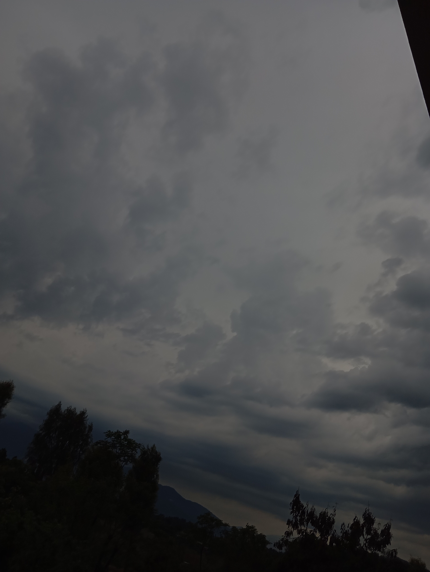 The image shows a dark, cloudy sky over a landscape in Lassan, Pakistan. Trees are silhouetted in the foreground, suggesting a forested or park area. A mountain is visible in the background. The overall scene conveys a sense of gloom or impending rain. No people are visible.