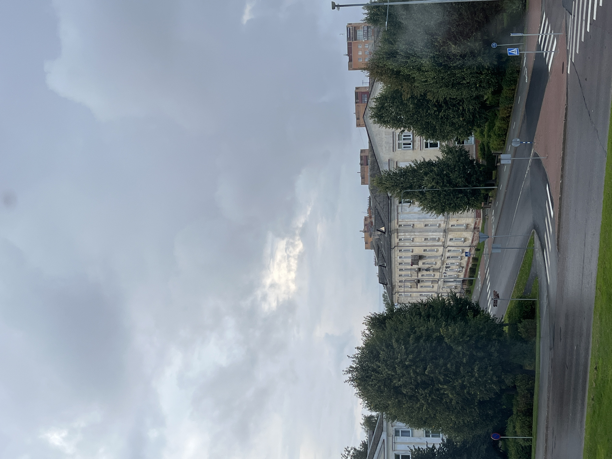The image shows a street view in Kohtla-Järve linn, Estonia. The sky is overcast with grey clouds. In the foreground, there is a wet asphalt road with pedestrian crossings. A large, mature tree with dark green foliage is prominent on the left side of the road. Behind the tree, a light-colored building with multiple windows and a dark roof is visible. Across the street, a long, light-beige building with a series of windows, balconies, and a dark roof stretches into the distance. Further back, other buildings, some taller with red-brick exteriors, are visible. Street signs and lampposts are present along the road. The overall impression is of a cloudy day in a residential or mixed-use urban area.