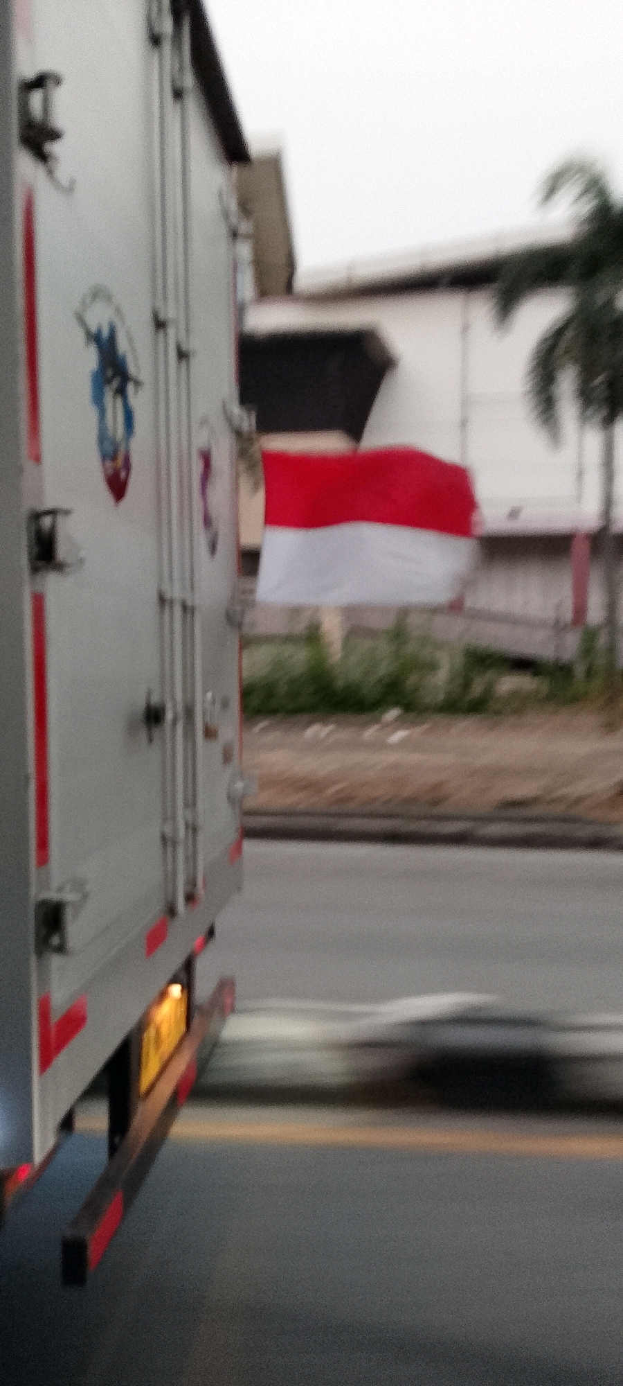 The image shows the side of a white truck, possibly a delivery vehicle, with red accents. The truck has a logo on its side, which appears to be a blue figure on a shield. Further back, an Indonesian flag, red and white, is visible attached to a pole or building. The truck is in motion, indicated by the motion blur. The background shows buildings and some greenery, suggesting an urban or suburban environment. The lighting suggests it is daytime. The license plate of the truck is visible, and it is yellow. The image is taken from a low angle, looking upwards. It is difficult to ascertain the exact location within Depok, Indonesia, without further context or landmarks.