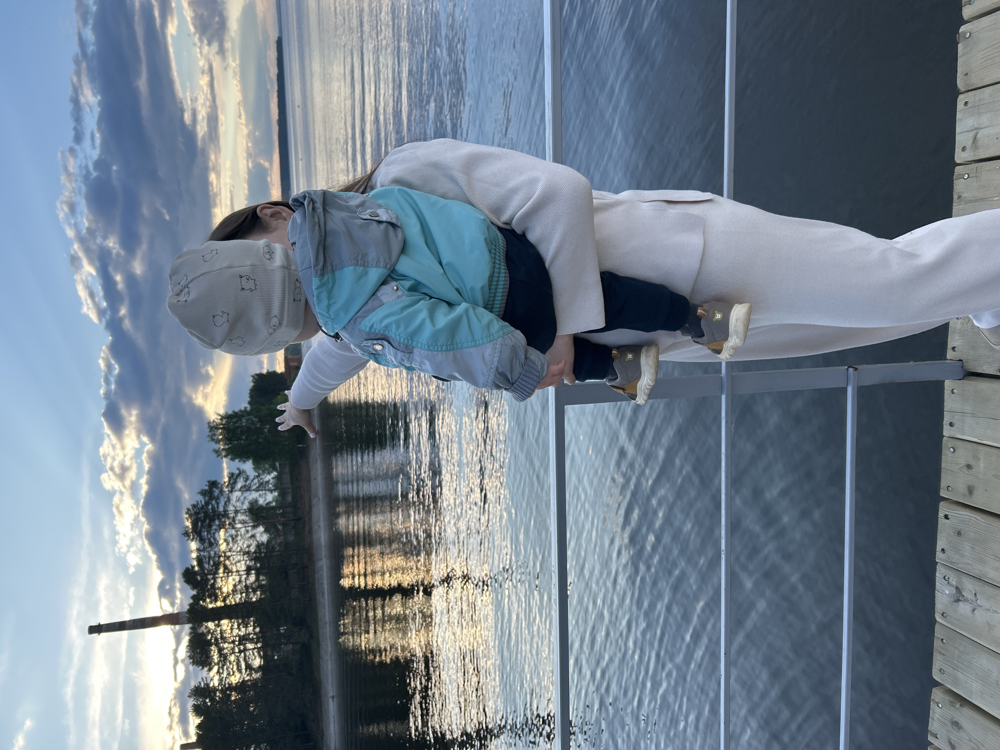 A mother is holding a young child in her arms near a body of water, specifically on a dock or pier. The child is pointing at something in the distance, possibly the horizon or some structure on the shore. The environment suggests it is a natural setting with trees and possibly a smokestack visible. The location appears to be in or near Chaikovsky, Russia, based on the presence of industrial infrastructure near the water. The time of day is likely near sunset, given the lighting conditions in the sky and the reflections in the water.