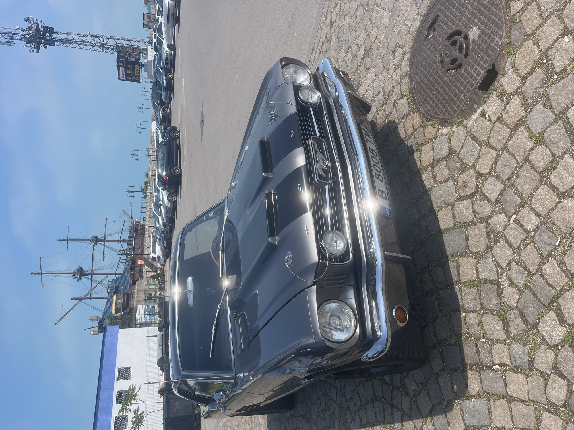 This image captures a vintage, dark grey Ford Mustang convertible, likely a first-generation model from the 1960s, parked on a cobblestone surface. The Mustang is detailed with distinctive silver racing stripes on its hood, a chrome grille featuring the iconic horse emblem, and shiny chrome bumpers and trim. Its Bulgarian license plate, displaying "B 8008 TM" and the "BG" country code, is prominently visible on the front.
Adjacent to the car, a circular, heavily textured metal manhole cover is visible on the cobblestones, showing signs of wear and a small section of damage.
In the background, a line of modern vehicles is parked on an asphalt road. Beyond these cars, a large, multi-masted sailing ship, reminiscent of an old galleon, is docked, indicating a port or marina environment. Further back, a tall metal truss structure, possibly a communication tower or part of a crane, rises against a clear blue sky. A small, distant sign on this structure shows some barely legible text, including "1500m." Several buildings, one featuring a white facade with blue accents, are also part of the backdrop.
The scene is brightly lit by strong daylight, evident from the sharp shadows cast by the car and other objects, suggesting a midday or early afternoon setting with clear, sunny weather. The combination of the Bulgarian license plate and the port setting, confirmed by the context, places the location in Varna, Bulgaria. No people are visible in the image.