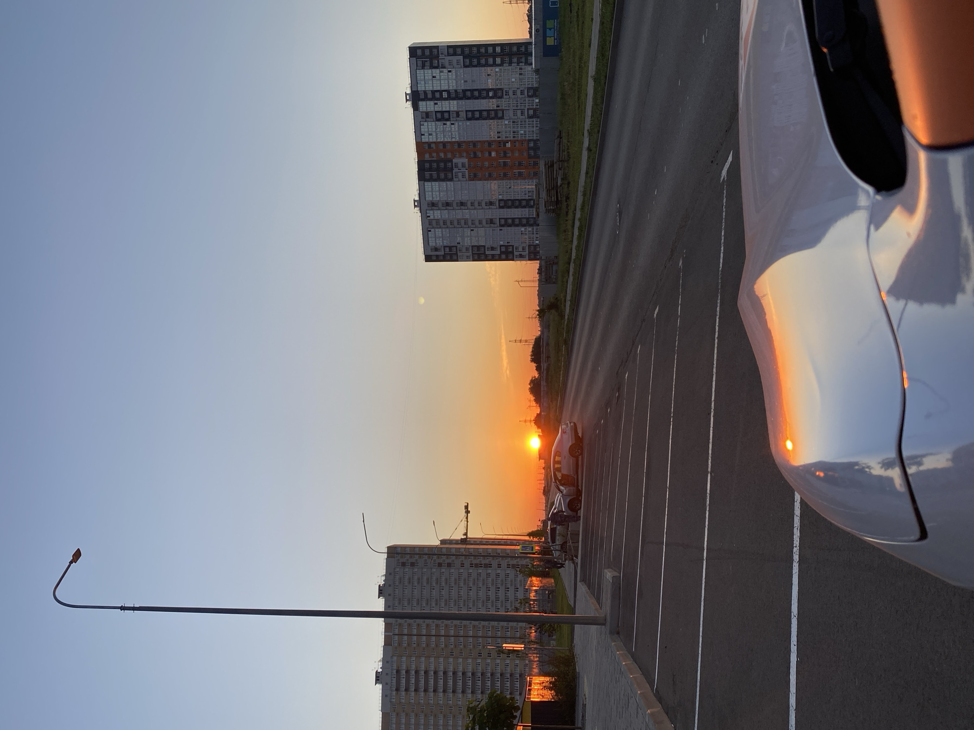 This is a photograph taken from a street-level perspective in an urban environment at sunset. In the foreground, the hood of a white car is visible, reflecting the warm hues of the setting sun. The car is parked in a paved parking lot with visible white parking lines. Beyond the parking lot, a paved road runs horizontally across the frame.
On the left side of the image, a tall, modern apartment building with many windows dominates the mid-ground. Adjacent to it, a sidewalk and a few trees are present. A lamppost stands prominently to the left of the apartment building, extending into the sky. In the background, another tall, modern apartment building with a distinctive facade is visible.
The sky is a gradient of pale blue at the top, transitioning to a vibrant orange near the horizon where the sun is setting. The sun itself is a bright, concentrated disc of light. Power lines and utility poles are visible in the distance, suggesting a developed area. Two cars are parked on the side of the road near the first apartment building, one of which has its doors open. The overall scene depicts a quiet moment in an urban setting during sunset.