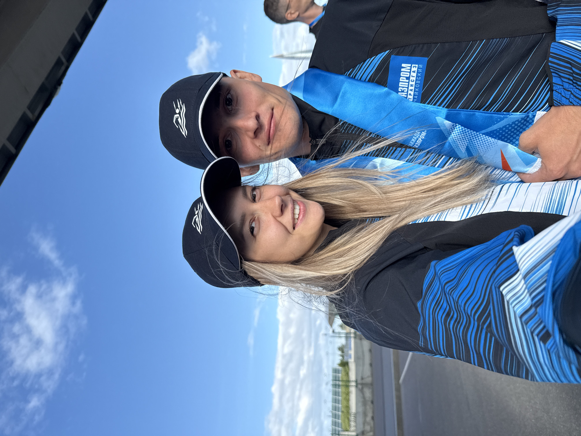 Two people, a woman and a man, pose for a selfie outdoors under a bright blue sky with some clouds. They are both wearing black baseball caps with a white logo depicting a swimmer. The woman, on the left, has long blonde hair and is smiling. The man, on the right, has short brown hair and a neutral expression. Both are wearing black jackets with blue and white abstract designs. They both have blue lanyards with medals around their necks, featuring the "Gazprom Transgas Chaykovsky" logo. The background shows an outdoor paved area, some fencing, and a glimpse of a building structure above. The location is not identifiable as Saint Petersburg.