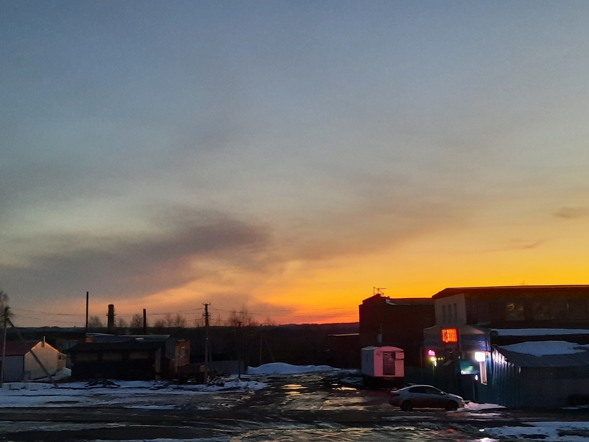 The image depicts an urban scene in Krasnogorskoye, Russia, at dusk. The sky transitions from blue-gray to orange as the sun sets. In the foreground, the ground is covered with snow and puddles reflecting the light. A car is parked near buildings and structures of varying sizes and designs. Some of the buildings have lights illuminated. Chimneys and power lines are visible on the horizon.