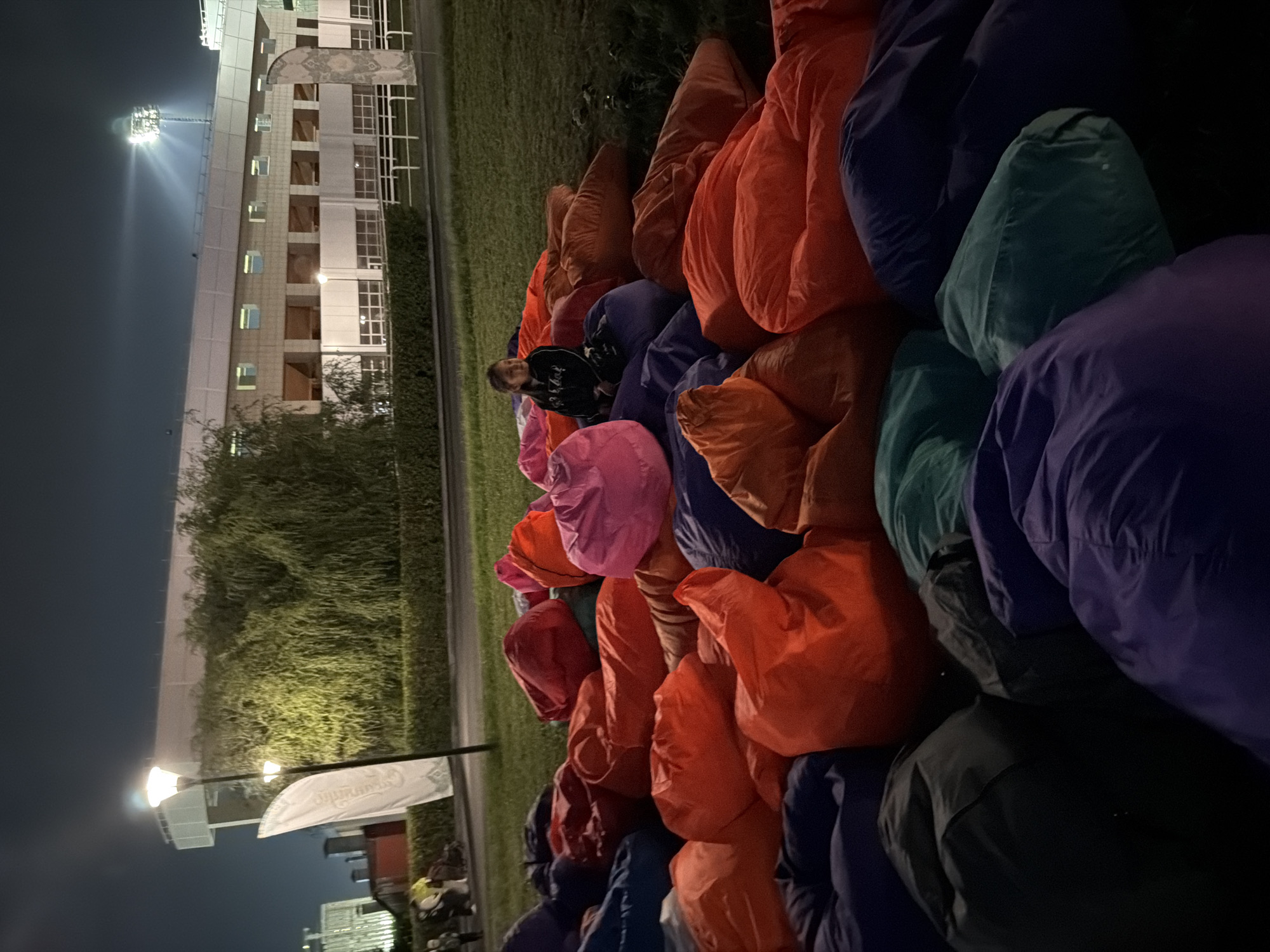 At night, in the city of Unknown, Unknown, a large collection of colorful bean bag chairs is spread across a grassy area. In the middle of the bean bag chairs, a young girl in a black hooded sweatshirt sits and looks to her left. In the background, there is a stadium with bright lights, a large weeping willow tree, and some other buildings.