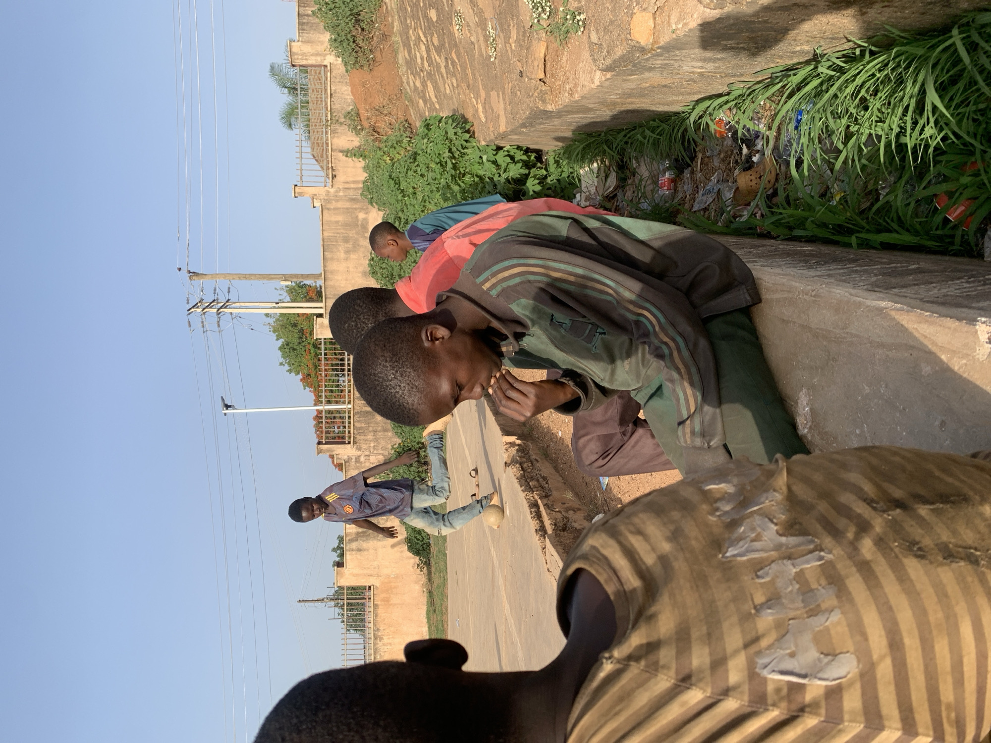 The image depicts a group of young people outdoors in Gura Top, Nigeria. Three individuals are leaning over the side of a concrete structure, possibly a culvert or drainage ditch, looking down into it. A fourth person is standing on a paved surface, wearing jeans and a short-sleeved shirt, with a soccer ball by his foot. Debris and vegetation are visible inside the concrete structure. The location appears to be an urban or semi-urban area, with buildings, walls, and utility poles in the background. The sky is clear and blue. A section of a person is visible in the foreground.