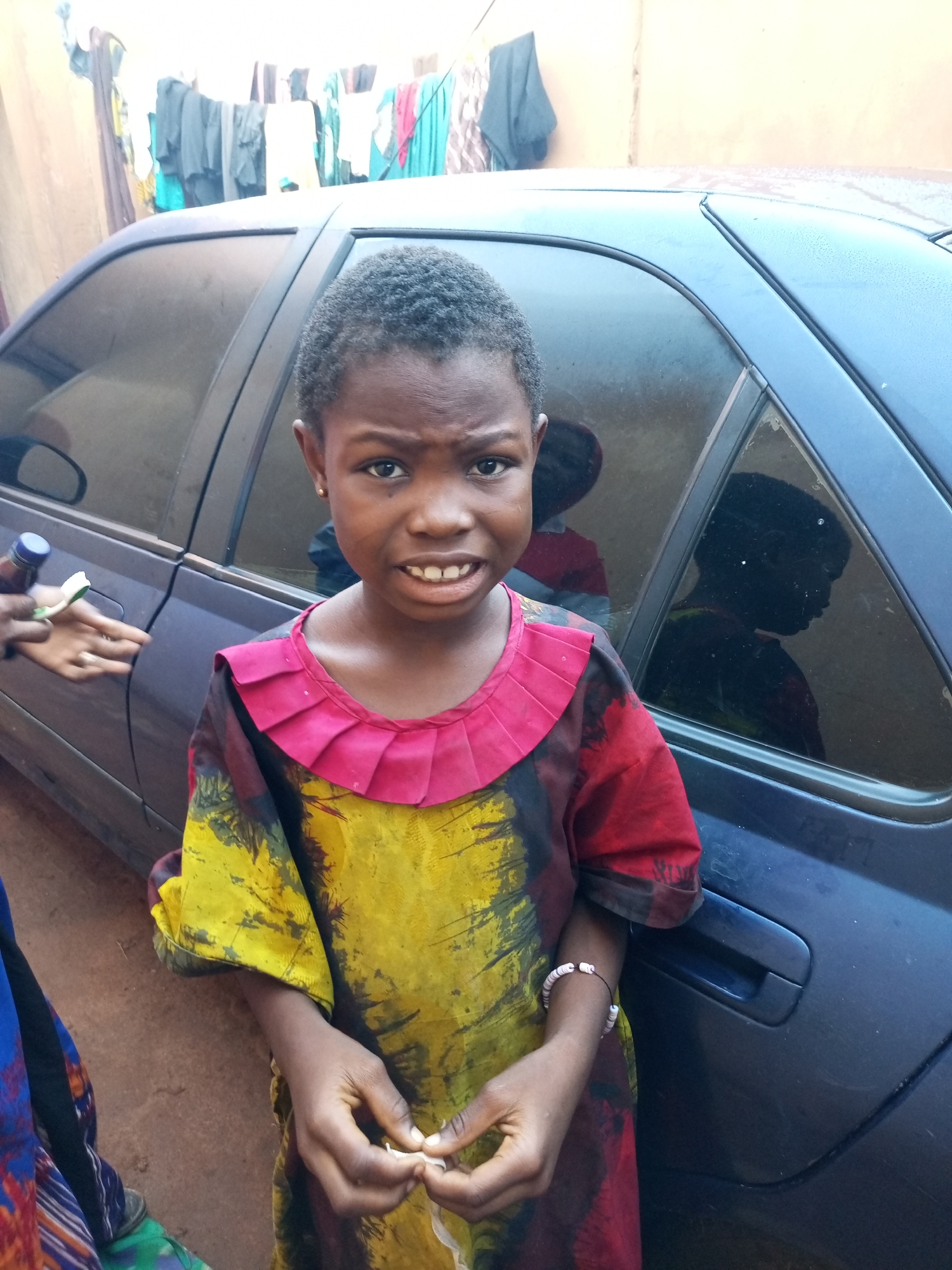 A young girl, appearing to be of African descent, stands in front of a dark blue car, likely in Tempure, Nigeria. She has short, dark hair and a slightly furrowed brow, her mouth open in a way that suggests she might be speaking or reacting to something. She is wearing a brightly colored, tie-dye dress with a pink ruffled collar. On her left wrist, she wears a bracelet made of white and pink beads. Her hands are clasped in front of her, with her fingers subtly manipulating something small and white. To the left of the frame, another hand is visible, holding a small bottle with a blue cap and a white object, possibly a toothbrush, suggesting a candid or interactive moment. In the background, laundry hangs on a line outside a building, indicating a residential setting. The car's windows are tinted, and a reflection of another person can be seen in the rear window.