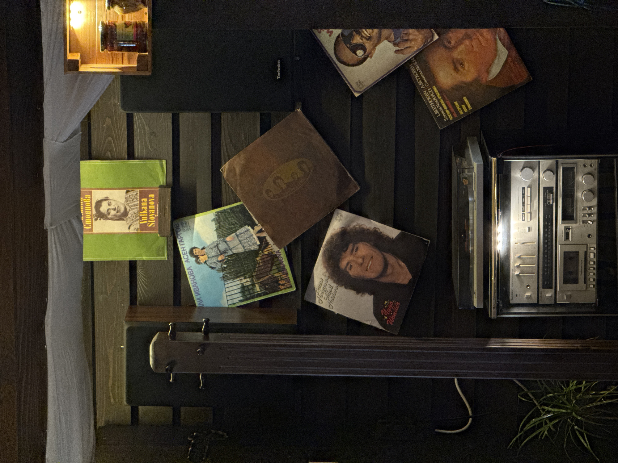 The image shows a close-up of a wall display, possibly in a bar or restaurant, consisting of vintage musical items and decorative objects.
The background appears to be made of dark wooden slats. Positioned on the wall are several vinyl record covers, most of them appear to be Bulgarian. The album covers are:
* "Vulkana Stoyanova" - Folk songs in the top left corner.
* "ILI IVANOVA ASEN GARGOV" - Located to the right of the previous album.
* "Lygo Wonder" - located below the previous album on the right side.
* "Sonatas Nos.8.14.23" - Top right corner.
A vintage Technics stereo component is visible on the right side.
Below that is part of the base of a bass instrument. The very top left corner is a wooden box with three glass jars. On the top left is a light colored curtain. The location appears to be in Sofia, Bulgaria due to the Bulgarian titles of the vinyls.