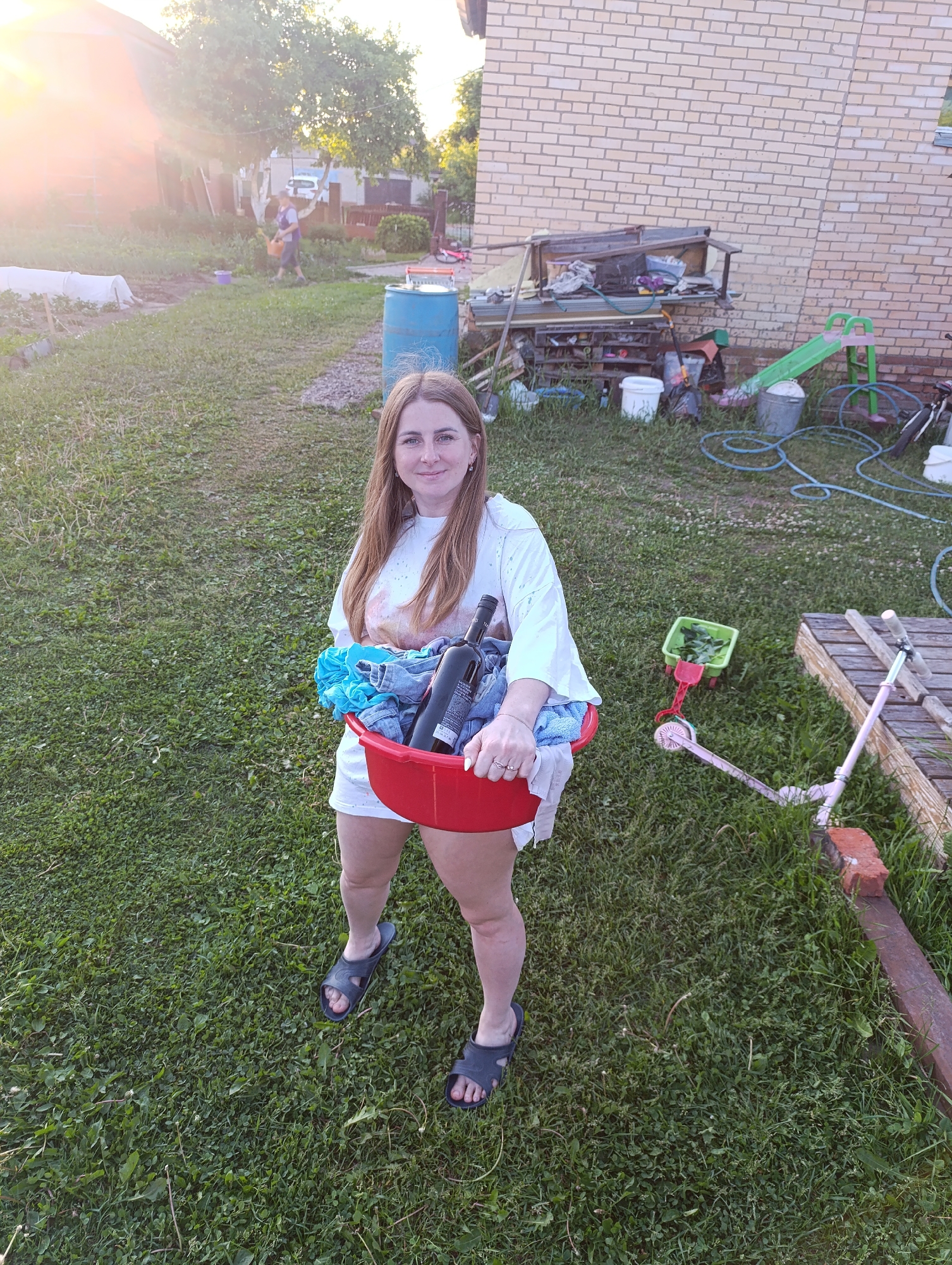 The image depicts an outdoor residential backyard scene in Unknown, Unknown, under what appears to be late afternoon or early evening sunlight, indicated by a prominent lens flare in the upper left and warm, golden illumination.
In the foreground, a light-skinned adult female stands facing the camera, smiling. She has long, light brown hair and is wearing a white oversized t-shirt with a faint pink/orange design and white shorts. She wears dark blue slide sandals. She holds a red plastic basin with both hands, containing blue folded fabric (possibly a towel), several denim items (likely jeans), and two dark-colored bottles, consistent in shape with wine bottles, one with a partially visible white label. She has small earrings, a ring on her left ring finger, and her fingernails are painted white.
The ground is covered in green grass with patches of clover and weeds. To the right of the woman, a pink child's kick scooter and a small green plastic wagon filled with green leaves are present. Further right, against the light tan brick wall of a building, is a disorganized pile of various items including a green child's slide, a blue coiled garden hose, white and metallic buckets, and what appears to be shelving and other outdoor debris. A single red brick is visible near a wooden plank.
In the mid-background to the left, a light-skinned adult male walks towards the right, carrying a green watering can. He is wearing a t-shirt and shorts. Behind him are garden beds, some covered with white protective fabric, and a blue plastic barrel. A light-colored car is partially visible in the distance behind the man. Several trees with dense green foliage are distributed across the background, and another building is partially visible on the far left, obscured by sunlight.