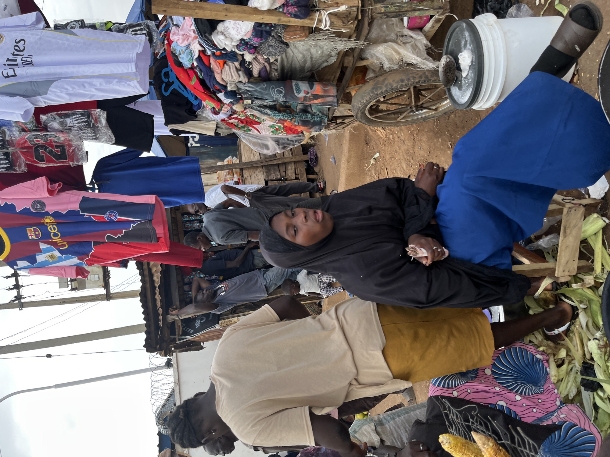 The image depicts a bustling street market scene in Rafin Par, Nigeria. In the foreground, a young woman wearing a black hijab and a long blue skirt sits beside a large white bucket, her hands clasped. To her left, a person wearing a beige t-shirt and a mustard-colored skirt stands, facing away from the camera, with a bundle of corn stalks visible. Behind them, several individuals are browsing at a stall displaying an array of colorful t-shirts, jerseys, and other clothing items hanging from a frame. The market appears to be outdoors, with a makeshift structure overhead and a dirt ground. The overall impression is one of vibrant commerce and everyday life in a Nigerian city.