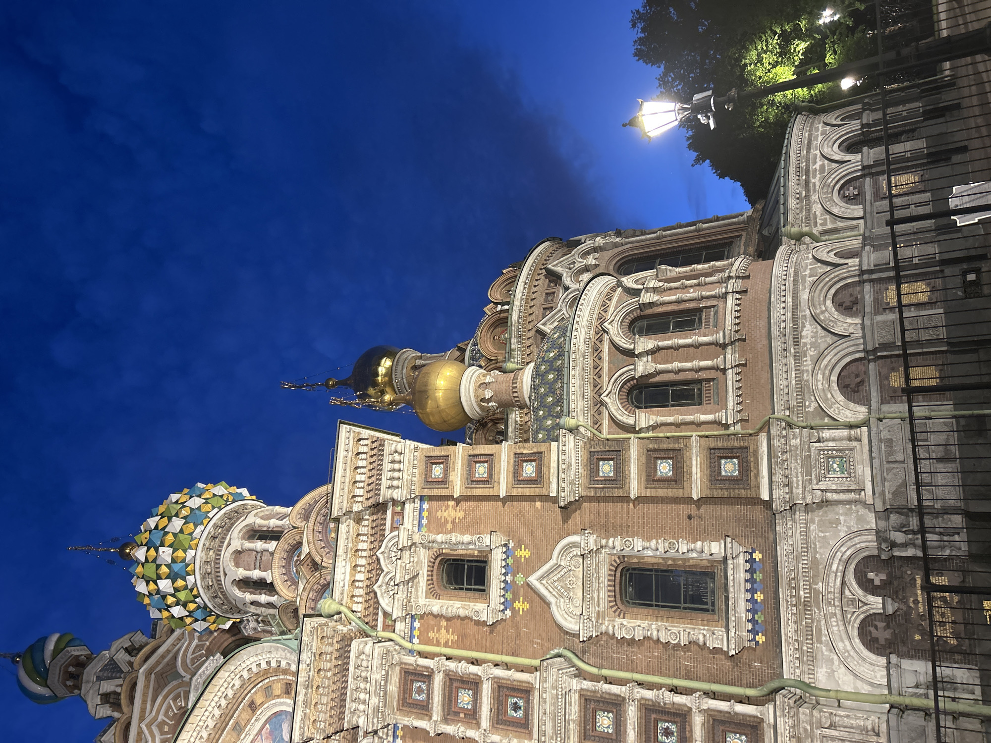The image shows the Church of the Savior on Spilled Blood in Saint Petersburg, Russia. The ornate Russian Revival style church features distinctive onion domes, colorful mosaic details, and brickwork. The sky is a deep, dark blue, indicating it is either dusk or nighttime. A vintage lamppost with a glowing light is visible in the foreground, illuminating part of the building and surrounding trees. A black metal fence runs along the bottom of the frame, separating the viewer from the church. The overall impression is of a beautifully illuminated architectural landmark at twilight.