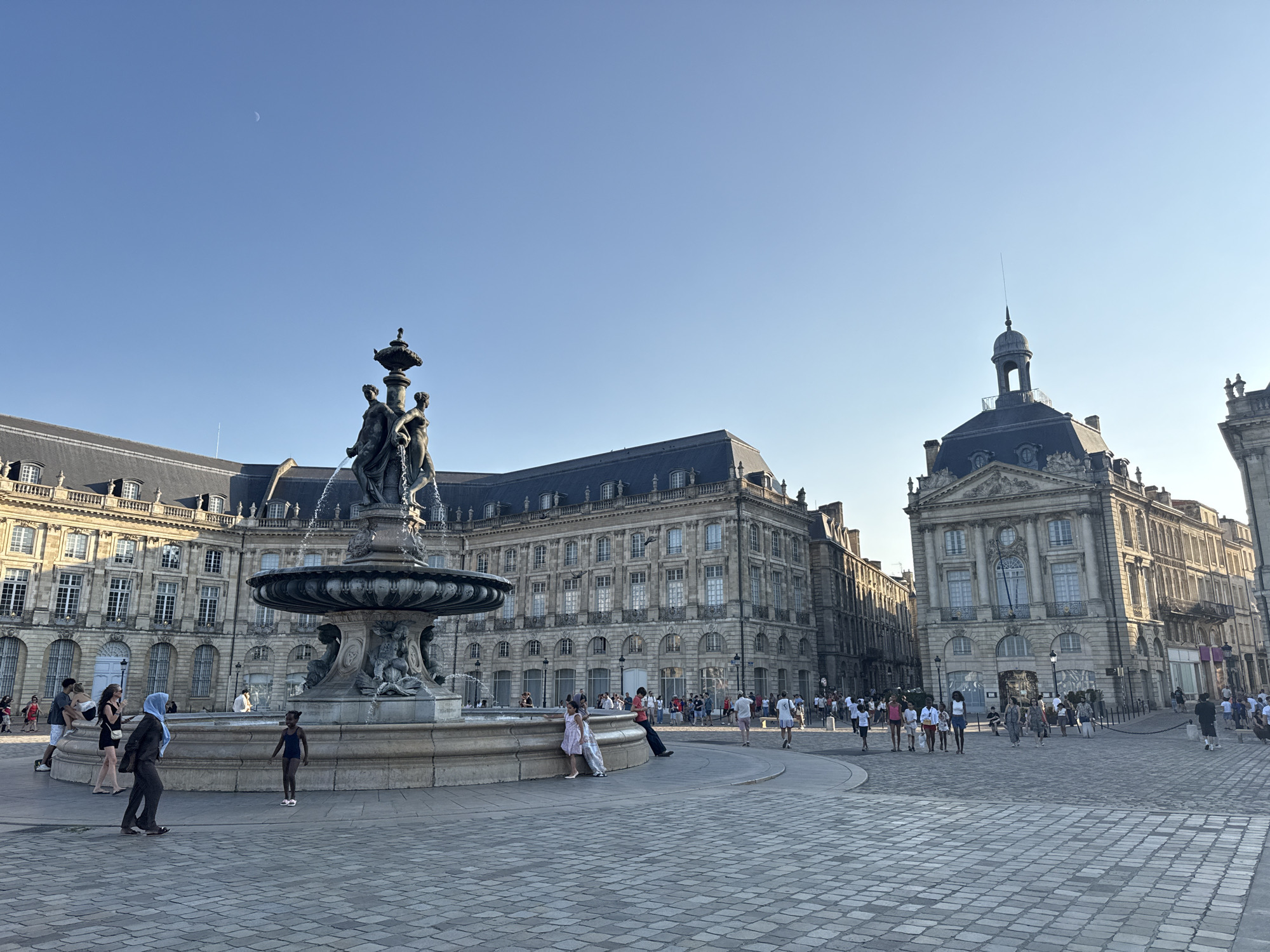 The image captures a wide-angle view of the Place de la Bourse in Bordeaux, France, under a clear blue sky with a crescent moon visible. In the foreground, a large, ornate fountain with sculpted figures is the primary focus. The fountain is surrounded by a paved plaza with cobblestone patterns. A diverse group of people are scattered throughout the scene, some walking, some standing, and some sitting near the fountain.
To the left and behind the fountain, a grand, classical-style building with multiple stories and arched windows stretches across the frame. Its facade is detailed with architectural ornamentation and is bathed in sunlight. To the right, another equally impressive building with a domed structure and classical detailing stands. This building also features numerous windows and is a prominent feature of the cityscape.
The people in the image are dressed in casual summer attire, suggesting a warm day. A young girl in a dark swimsuit stands near the fountain, while other individuals and small groups are seen strolling across the plaza. The overall atmosphere appears to be one of leisure and sightseeing. The lighting suggests it is late afternoon or early evening, given the long shadows cast by the buildings. The iconic architecture of the Place de la Bourse, the presence of the fountain, and the clear sky are all characteristic elements of this well-known Bordeaux landmark.