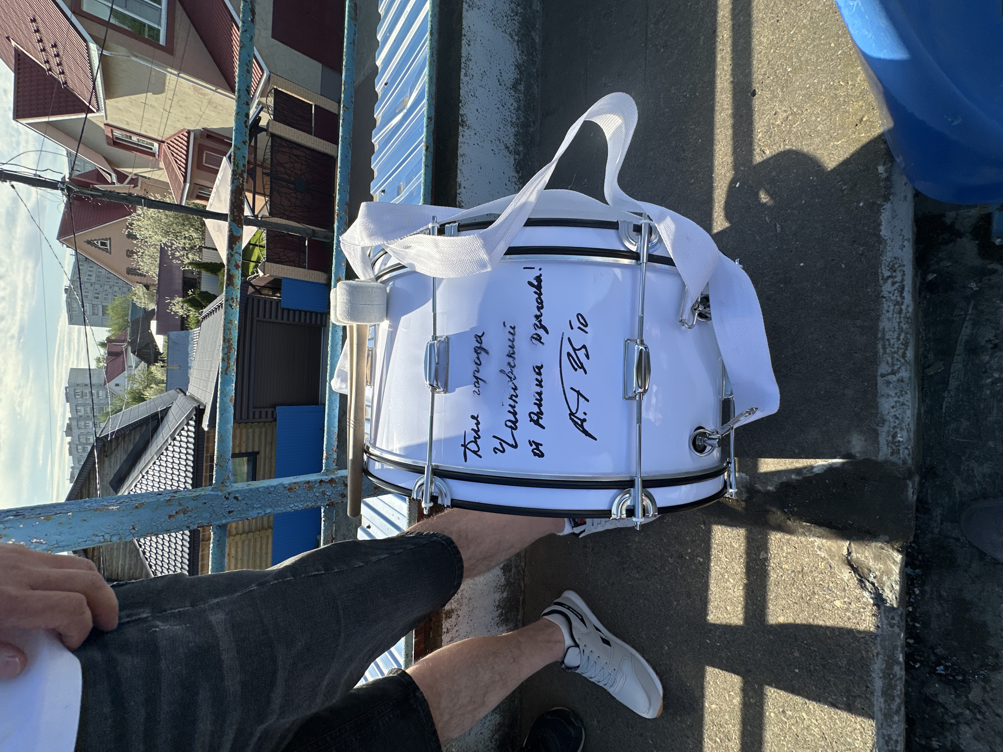A white drum with black trim is positioned on an elevated concrete surface, likely an overpass or bridge, with the legs of a person visible in the foreground. The drum has a white strap and drumstick holder attached. Black handwritten text is visible on the drum's surface. The person is wearing dark gray shorts and white sneakers. A blue metal railing is in the background to the left. Beyond the railing, residential buildings and trees are visible, indicating an urban or suburban setting. The location is the city of Chaikovsky, Russia.