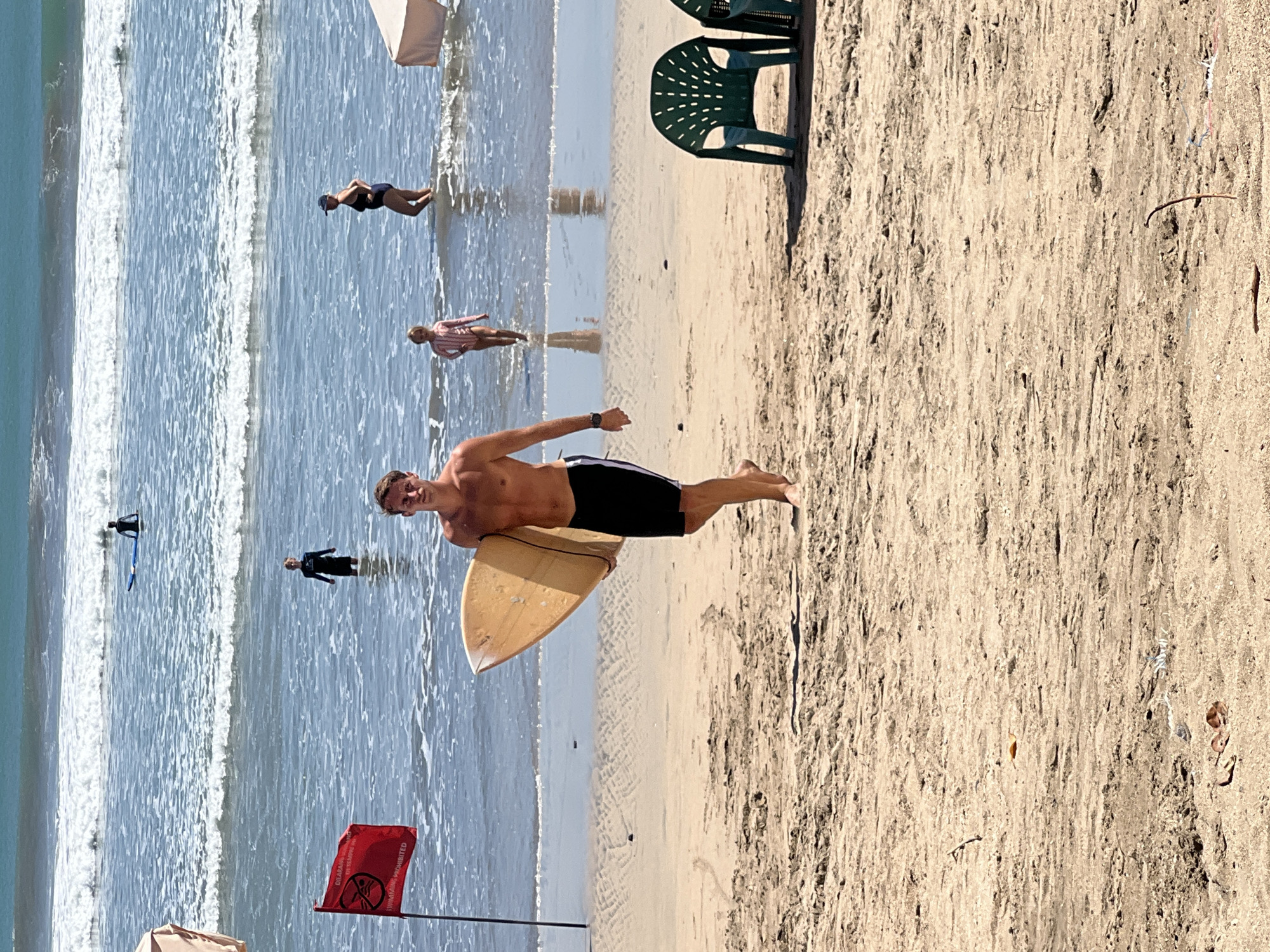 In Kuta, Bali, Indonesia, on a sunny day at the beach, a young man with a surfboard walks towards the camera on the sandy shore. He is shirtless and wearing black swim trunks. In the background, several people are visible in the ocean. A red flag, indicating "SWIMMING PROHIBITED," is planted in the sand to the left of the frame. The ocean waves are moderately sized, and the water is a clear blue. Two green plastic beach chairs are positioned on the sand to the right of the frame. The sand on the beach is a light brown color, with some debris and footprints visible. The sky is clear and blue, suggesting fair weather.