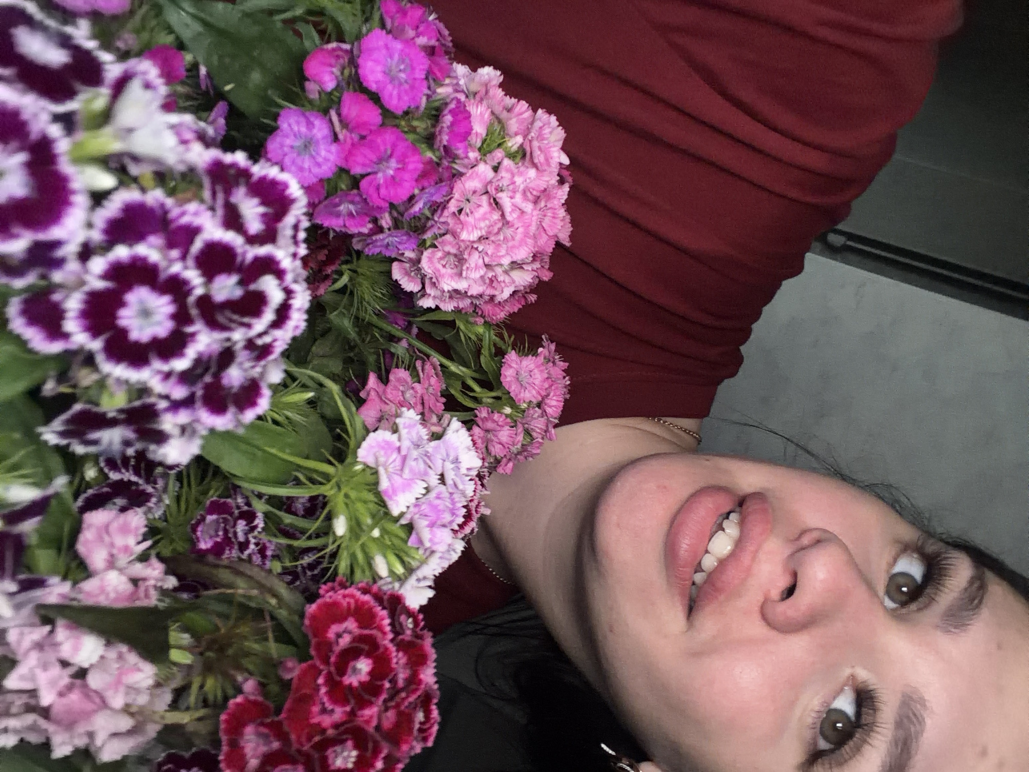 A close-up, overhead shot features a woman holding a bouquet of various pink and purple carnations. She is wearing a dark red shirt and her left arm is visible, partially obscured by the flowers. The woman has long, dark hair, prominent eyelashes, and is looking upwards and to the right, with her mouth slightly open. A gold chain necklace is visible around her neck. The background is a neutral gray wall. There is no indication of the city of Фоки, Russia, or any specific urban environment.