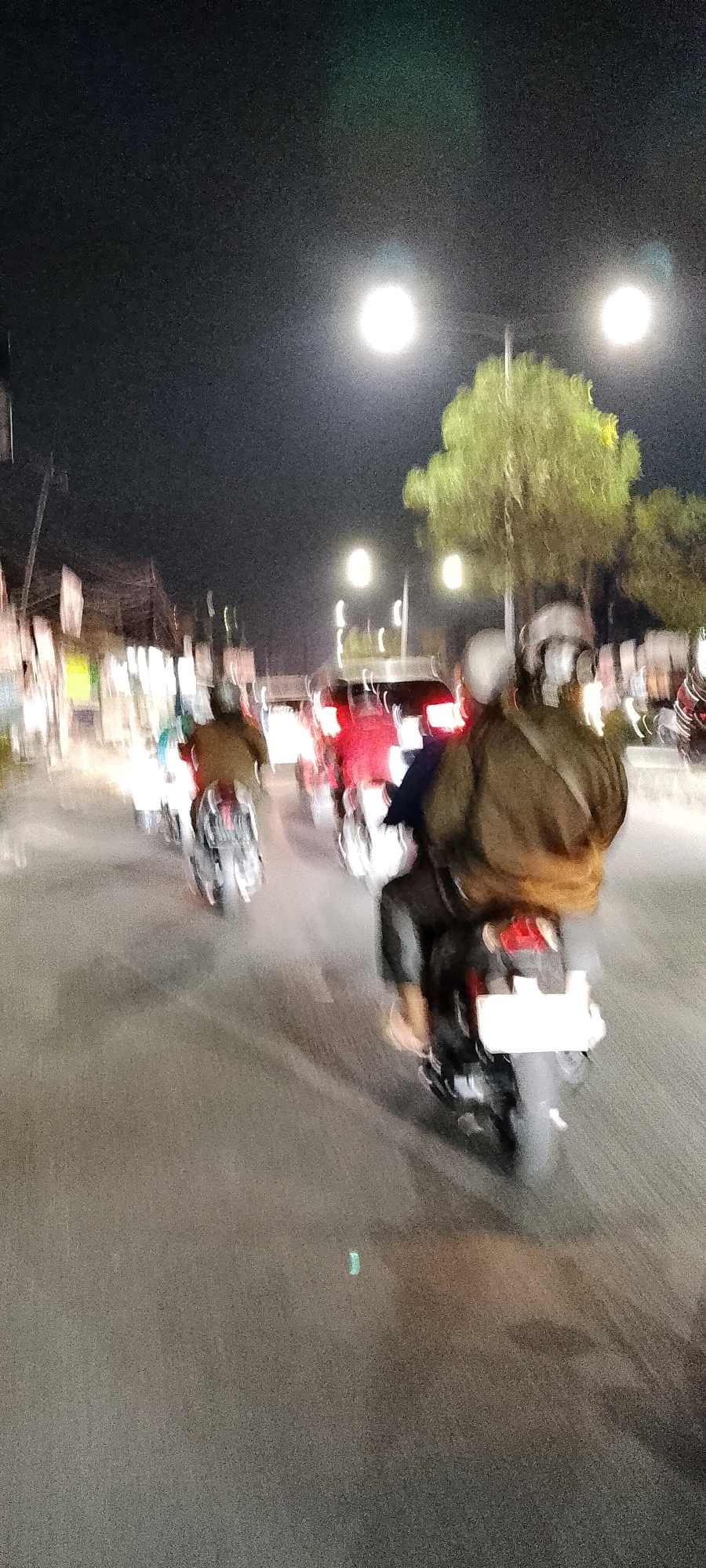 This is a night-time, blurry, low-light photograph of a street scene in Bekasi, Indonesia, possibly on Jalan Raya Chandrabhaga. The image captures several motorcycles in motion, indicated by motion blur. In the foreground, a motorcycle with two occupants is prominently visible. The rear passenger is wearing a dark jacket and a helmet, while the driver, partially obscured, is also wearing a helmet. The motorcycle has a white license plate. To the left and further down the road, another motorcycle with a single rider, wearing a brown jacket and a helmet, is also in motion. In the background, other vehicles and figures are suggested through streaks of light and indistinct shapes, some appearing to be other motorcycles and possibly cars. The street is paved and appears to be wet, reflecting the light from street lamps. On either side of the street, blurry structures that could be shops or buildings are visible, illuminated by light sources. Two bright streetlights are present in the upper portion of the image, casting a diffuse glow. A tree with green foliage is partially visible in the upper right quadrant, its leaves blurred by the motion. The overall impression is one of a busy, albeit dimly lit, urban street at night.