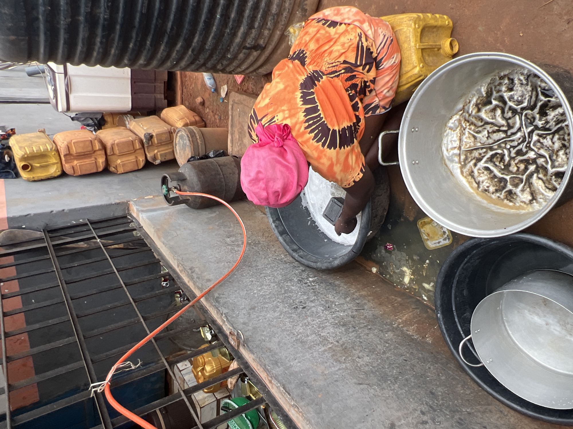 In Nkpor, Nigeria, a person wearing a bright pink headscarf and an orange and brown patterned top is washing items in a large black basin filled with white liquid. They are kneeling on a yellow jerrycan. Next to the basin, another large metal pot contains a dark, textured substance. In the background, several stacked jerrycans, a propane tank, and a large black ribbed water tank are visible, along with a washing machine. The scene appears to be outdoors, with a concrete wall and reddish-brown dirt ground. An orange hose runs from the left of the frame toward the basin.