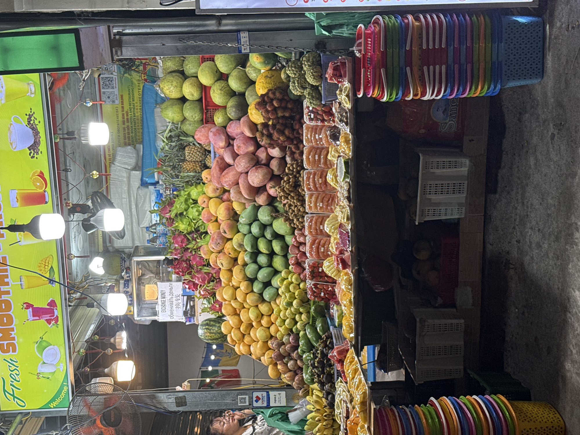 This image captures a vibrant fruit stand in Nha Trang Ward, Vietnam. A diverse array of fruits, including pomelos, rambutans, mangos, dragon fruit, and bananas, is prominently displayed. Many stacked, colorful plastic baskets are visible. A vendor is present. The stand has a bright yellow sign advertising "Fresh Smoothies." A sign indicating money exchange is also visible. A fan is present. The background appears to be a narrow street with limited lighting.