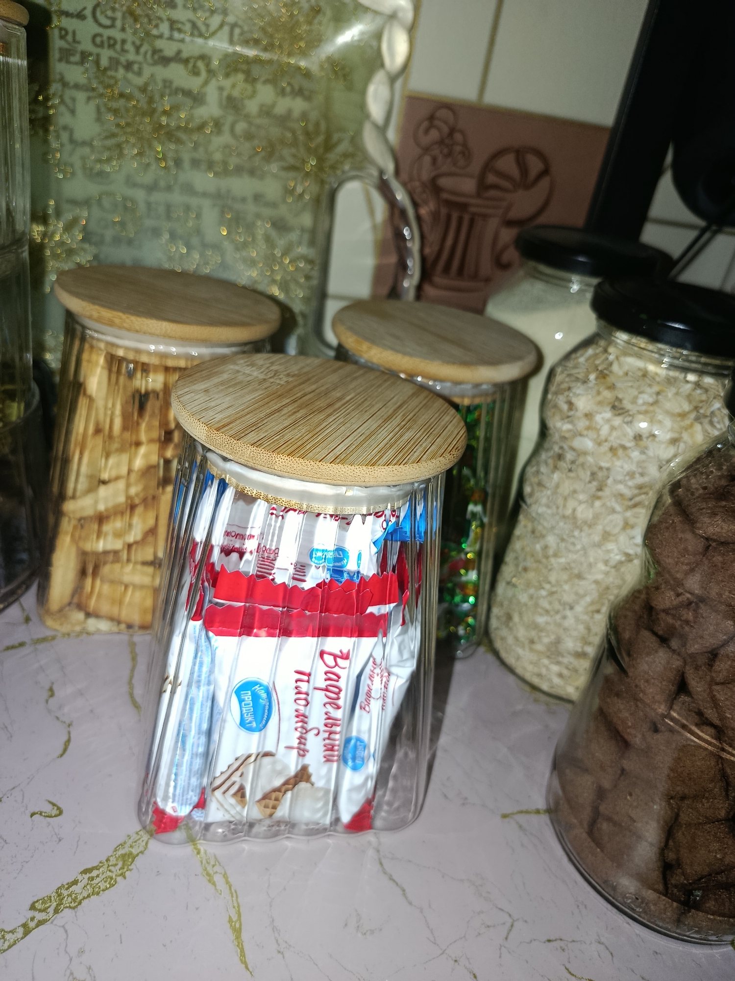 The image displays a collection of glass jars with bamboo lids, situated on a light-colored countertop with subtle green veining. In the foreground, a tall, clear glass jar contains individually wrapped wafer cookies with red and white packaging. The wrappers are decorated with text that appears to be in Cyrillic script and images of wafers. Behind this jar, another clear glass jar holds what looks like thin, dried plant material, possibly bamboo or wood, arranged vertically. To the right, two other glass jars are visible. One is partially filled with a light-colored granular substance, possibly oats. The other, on the far right, contains dark brown, irregularly shaped food items, potentially cookies or cereal pieces. In the background, a blurred green package with decorative golden elements is partially visible, along with a brown decorative tile featuring an embossed image of a cup. The lighting in the image is somewhat dim, creating soft shadows and highlighting the textures of the objects. The scene suggests a kitchen or pantry setting, focused on organized food storage. There is no visible indication of the location being specifically in Tashkent, Uzbekistan.