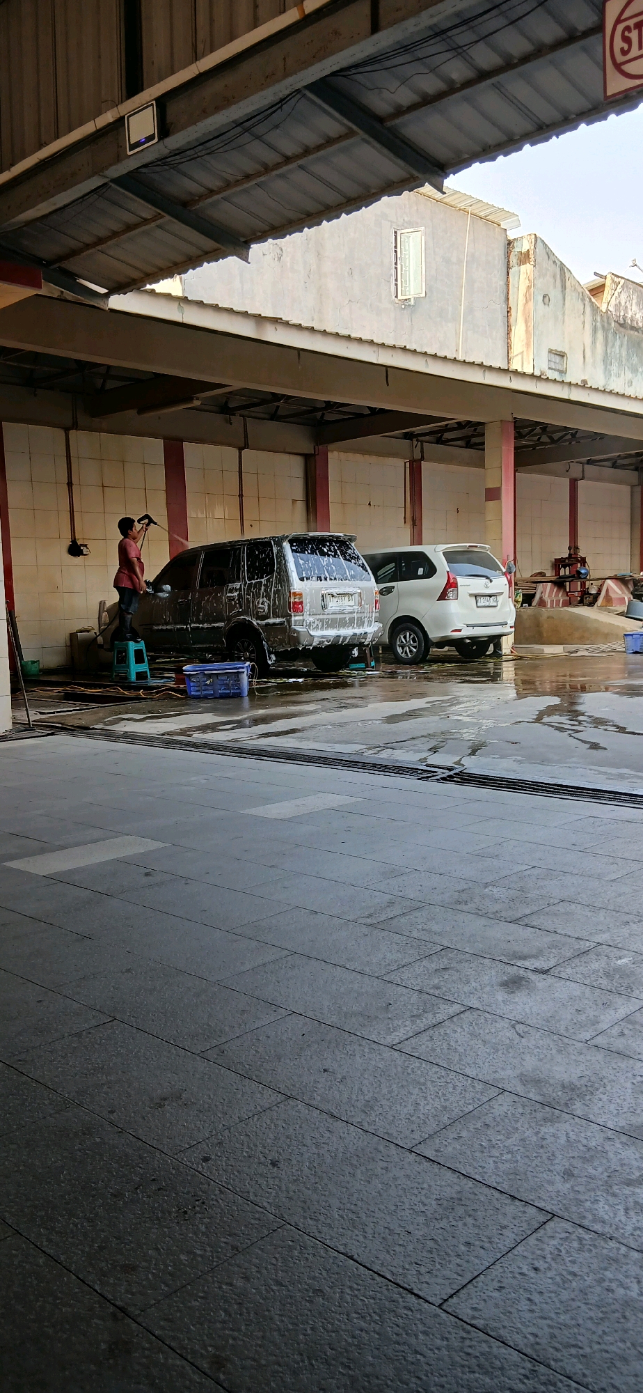 The image depicts an outdoor car wash facility in Samarinda, Indonesia, under a covered structure. In the foreground, a person wearing a red shirt and dark pants stands on a light blue stool, holding a spray nozzle and washing a silver SUV covered in white foam. The SUV is positioned in the center of the frame, and next to it is a white minivan, also partially covered in foam. The ground is wet and reflective, indicating recent washing. Various cleaning supplies, including blue crates and buckets, are visible around the vehicles. The background shows the interior of the car wash structure with tiled walls and a metal roof, as well as the surrounding urban environment with buildings. The scene captures the mundane activity of car washing in a typical Indonesian city.