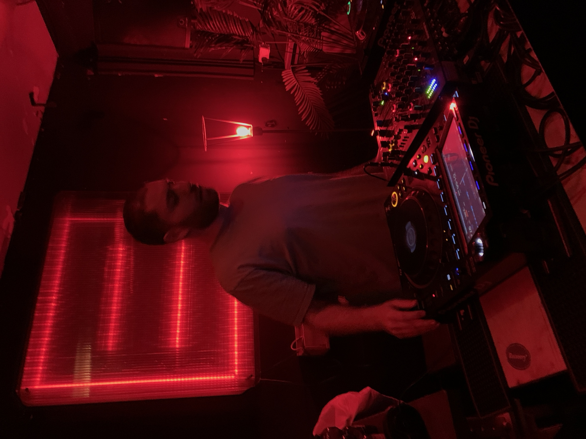 The image depicts a DJ working in a dimly lit setting, possibly a nightclub or a bar. The DJ, a man with short hair and a beard, is positioned behind a Pioneer DJ setup, which includes a mixer and CDJs. Red lighting dominates the scene, casting a strong hue over the equipment, the DJ, and the surrounding environment. A square-shaped light installation emitting red light is visible in the background. The presence of palm fronds suggests a decorative element. The overall atmosphere seems to be consistent with a nightlife scene in Tel-Aviv.