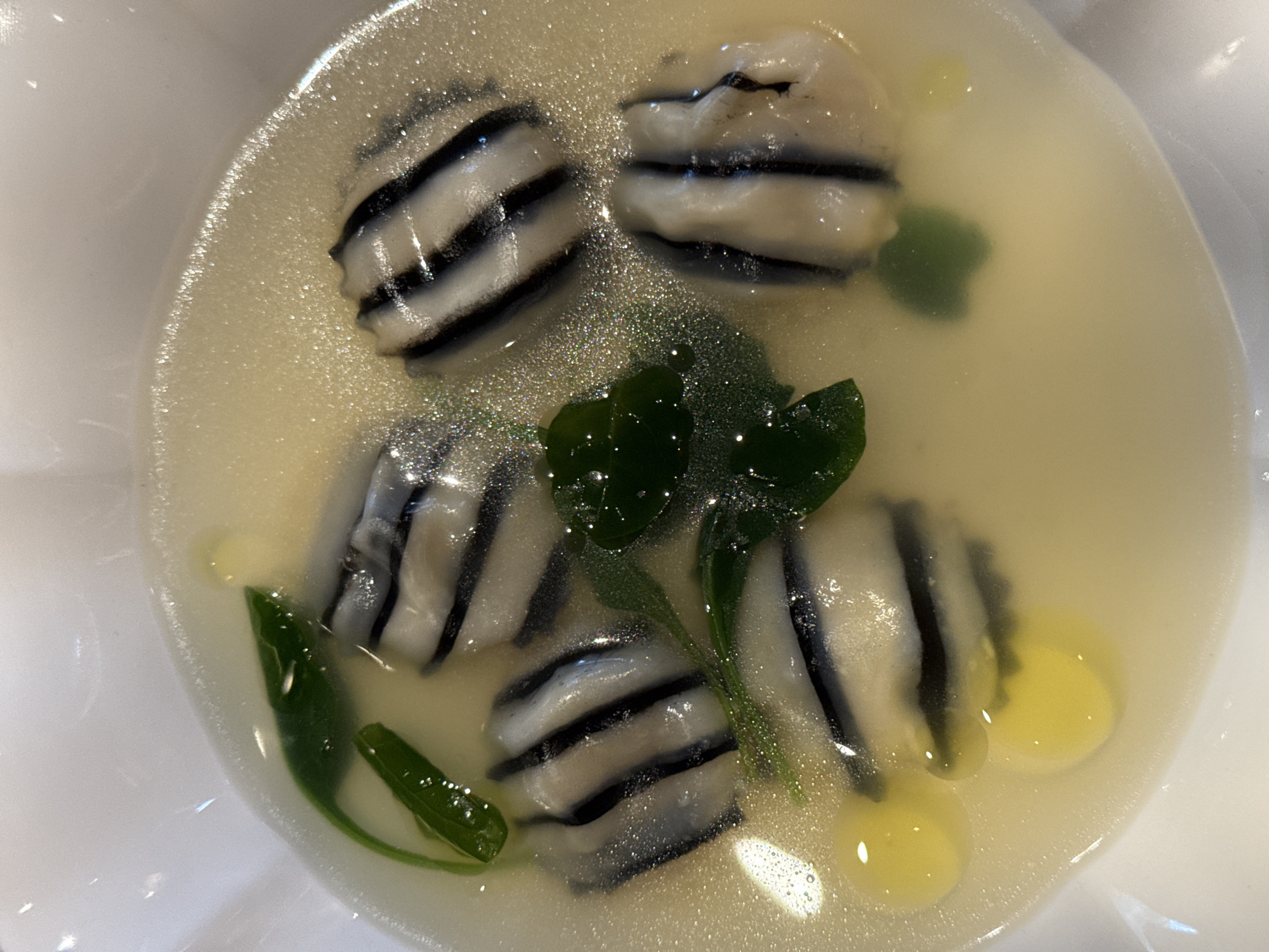 A close-up overhead shot shows a serving of food in a white bowl. The dish consists of several pieces of striped dumplings, alternating black and white, submerged in a light-colored broth. Green leaves are scattered among the dumplings, and small yellow droplets, likely oil, are visible on the surface of the broth. The background is the white rim of the bowl. The image does not contain any recognizable landmarks or elements that would allow for identification of its location in Nizhny Novgorod, Russia.