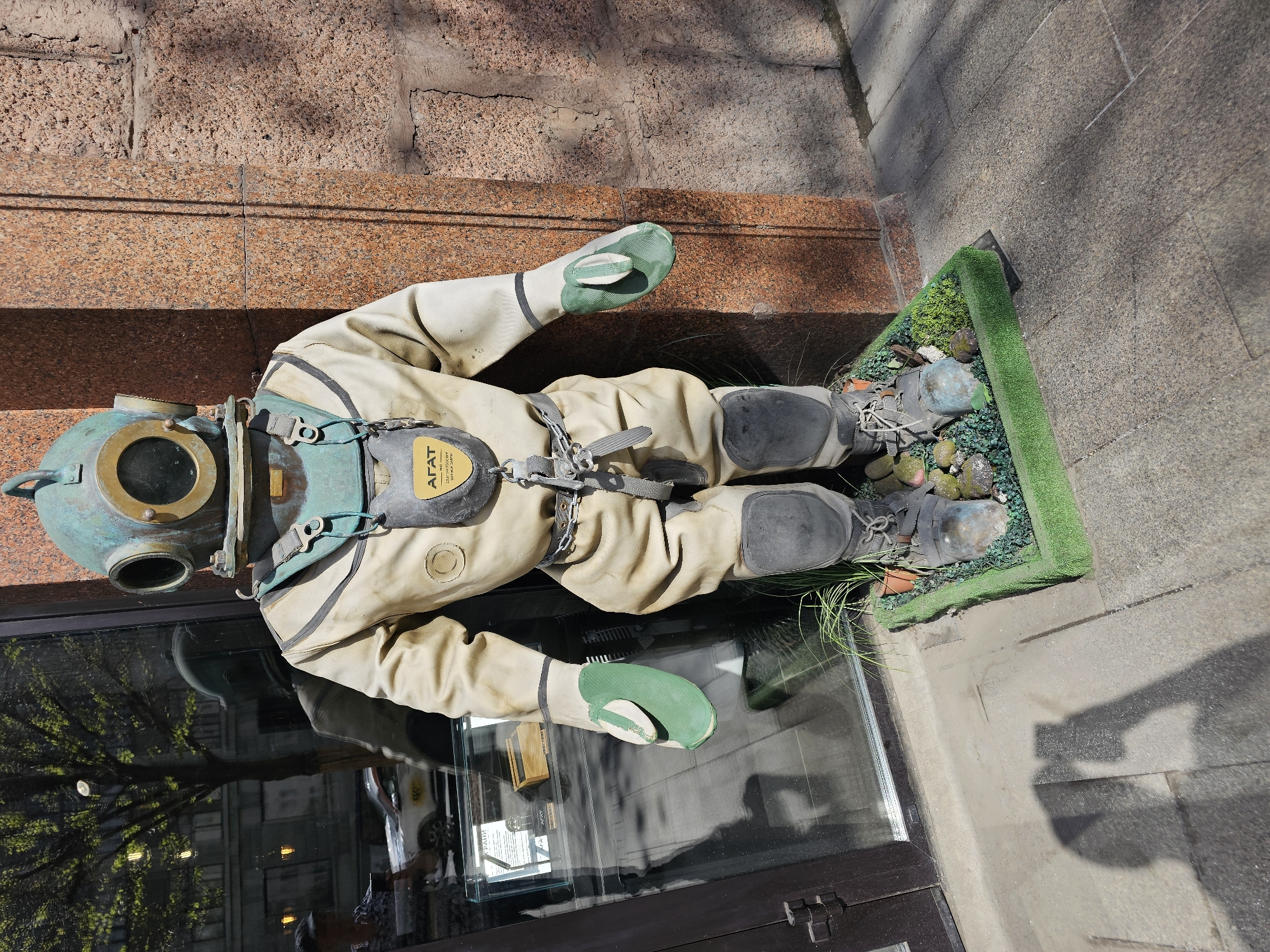 An antique diving suit is displayed on a street in Moscow, Russia. The diving suit has a bronze helmet and green gloves. The body of the suit is off-white. The suit lies on a small patch of green grass. The suit is positioned in front of a storefront window. A building facade is visible in the background.