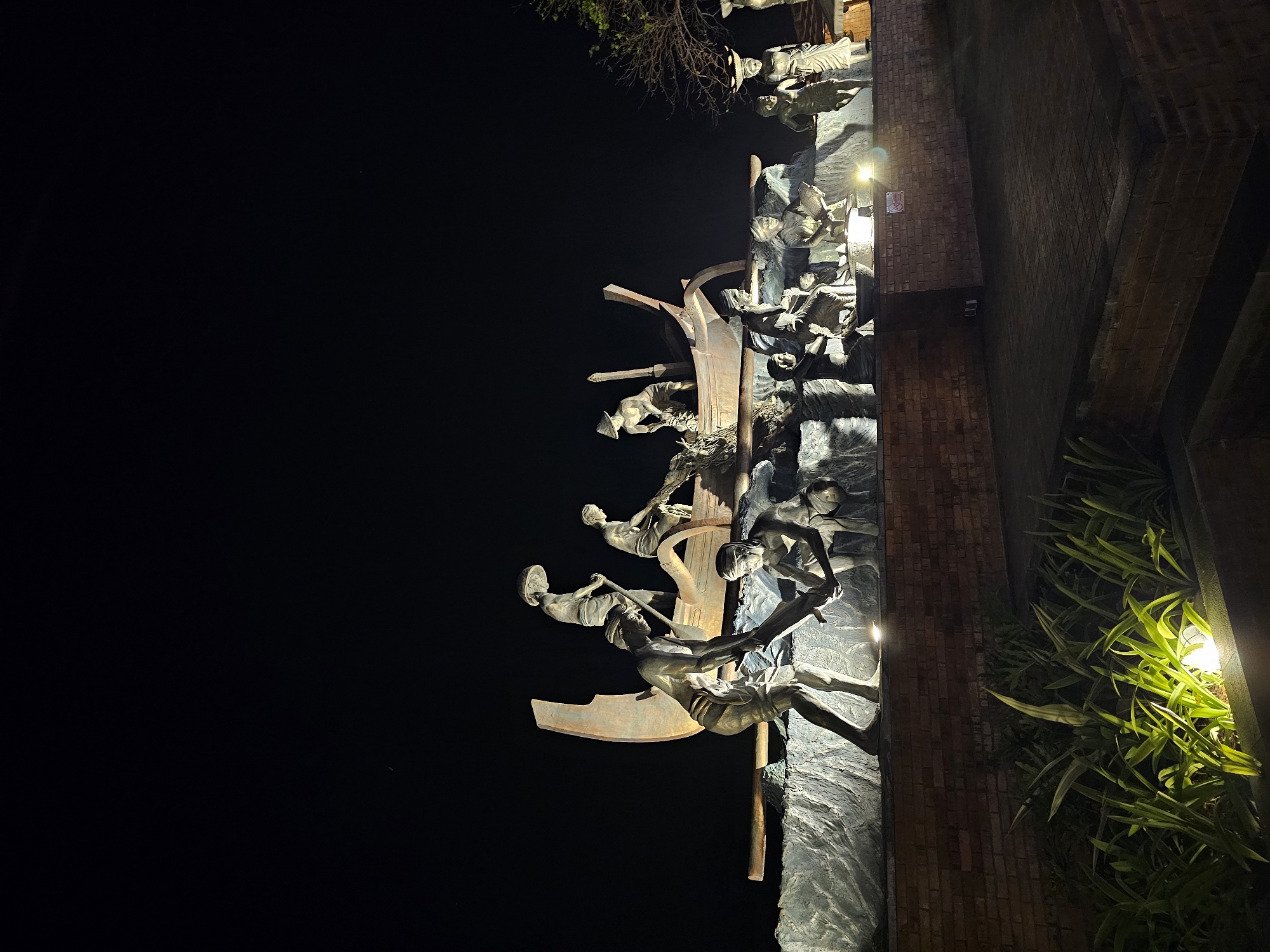 This is a night-time, low-angle photograph of a large bronze sculpture depicting a group of people working on a boat, likely engaged in fishing. The sculpture is illuminated from below, casting dramatic shadows. The figures are dressed in traditional clothing, including conical hats. The boat is positioned on a rough, textured base that simulates waves. In the foreground, there is a brick wall and lush green plants illuminated by ground lights. The background is a dark, clear night sky. This sculpture is located at the Monument Bajra Sandhi in Denpasar, Bali, Indonesia, which commemorates the struggles of the Balinese people.