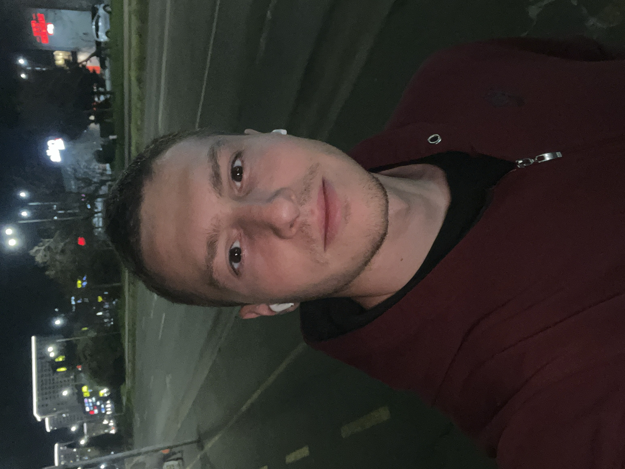 The image shows a man in a maroon jacket, wearing white earbuds, on a street in Burgas, Bulgaria at night. Streetlights and lit buildings are visible in the background.