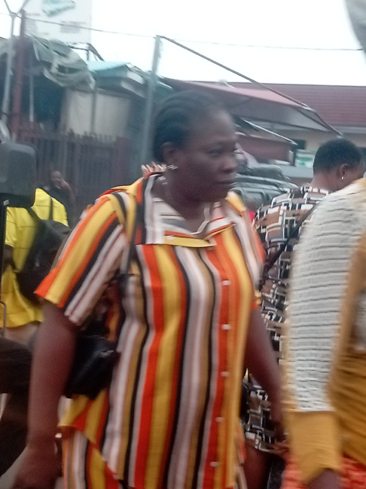 A woman in a striped shirt with a black purse is walking in what appears to be a busy street in Onitsha, Nigeria. Other people are nearby. There are buildings and structures visible in the background, including one with a billboard. The image is slightly blurry, and the lighting is somewhat overcast.