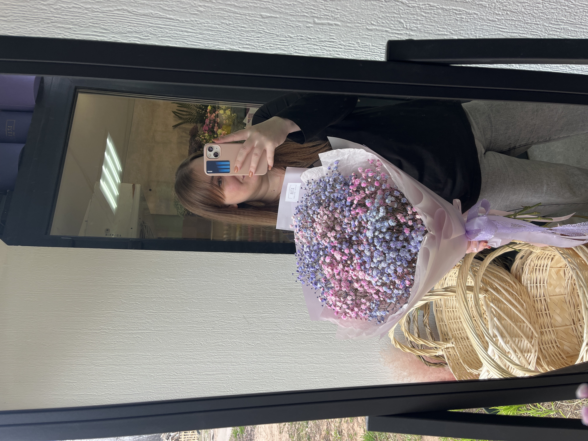 A reflection in a mirror shows a person holding a bouquet of pink and blue flowers in what appears to be a shop. The person is holding a pink mobile phone up to their face. Baskets are visible in the lower right corner, and other floral arrangements are seen in the background. The setting is likely a flower shop based on the objects and the type of photo. The location is speculated to be in Chaikovsky, Russia.
