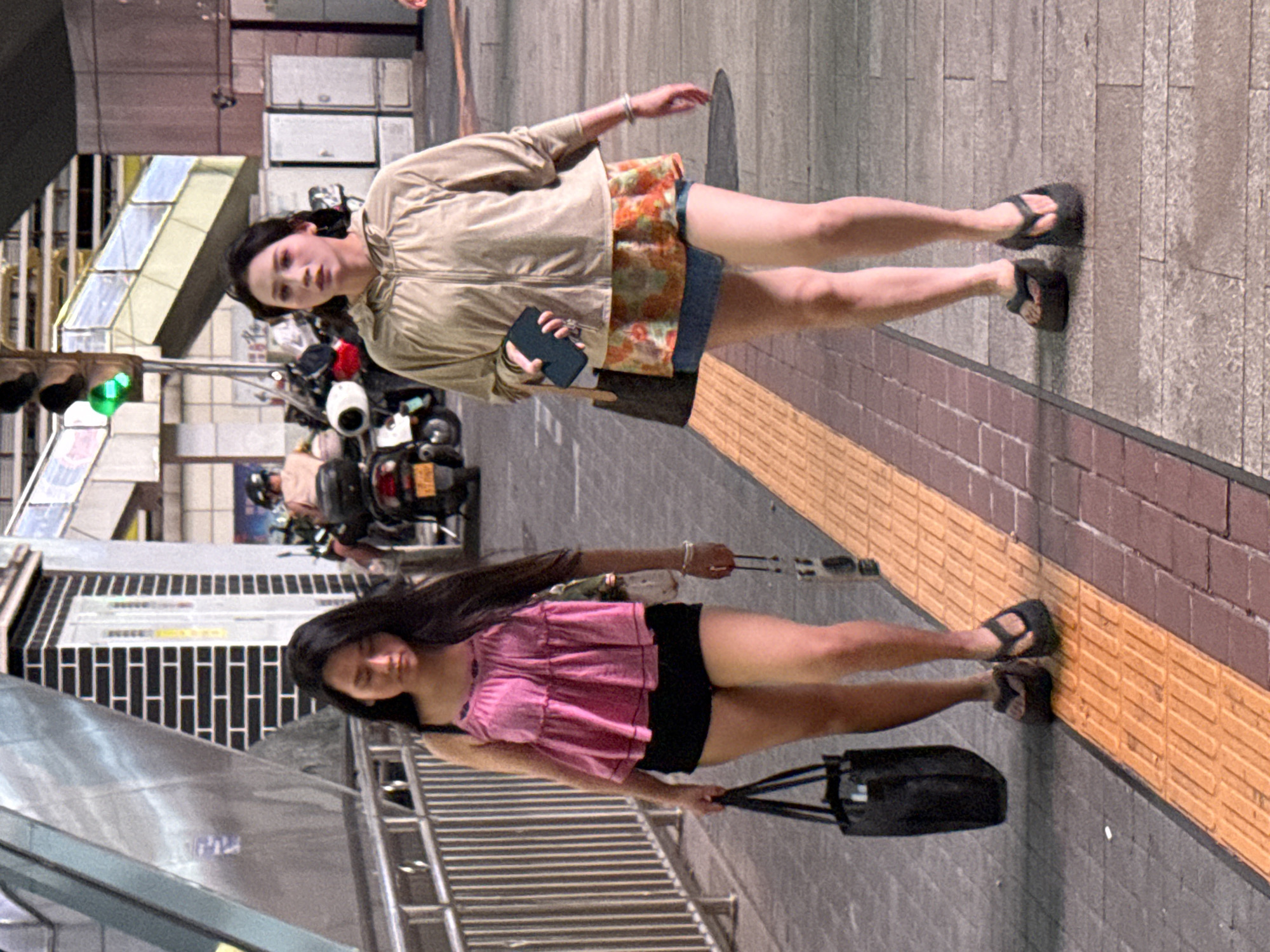 Two women are walking side-by-side on a sidewalk in Yuzhong District, China. The woman on the left is wearing a pink spaghetti strap top, black shorts, and dark flip-flops. She is carrying a black tote bag in her left hand and has sunglasses hanging from her right hand. The woman on the right is wearing a tan hooded jacket over a patterned skirt and denim shorts, and dark flip-flops. She is holding a black smartphone in her right hand and carrying a brown tote bag over her left shoulder. They are walking past a metal railing and a textured yellow and orange guideway. In the background, there are motorcycles and traffic lights.