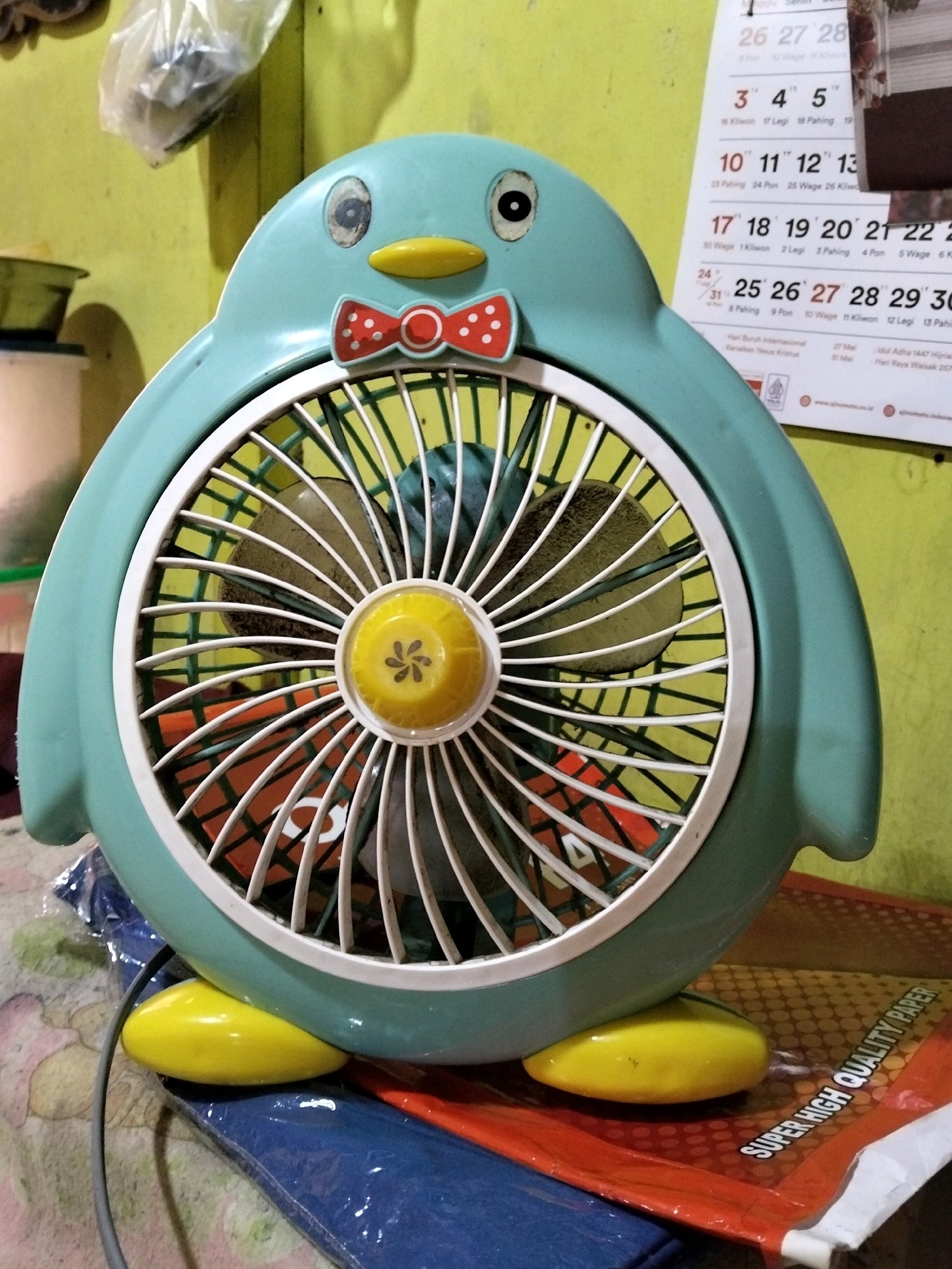 The image shows a teal-colored penguin-shaped fan on a surface covered with a blue and orange mat. The fan has a red and white polka dot bow tie, yellow beak, and black eyes. The fan blades are visible behind the cage, with a yellow circular center. In the background, a calendar with dates and Indonesian text is hanging on a pale yellow wall. The calendar appears to be from the year 2023. To the left of the fan, a plastic bag containing some objects is also visible against the wall. The lighting suggests an indoor environment. The specific location in Teluk Pambang, Indonesia, is not discernible from the visual information presented in the image.
