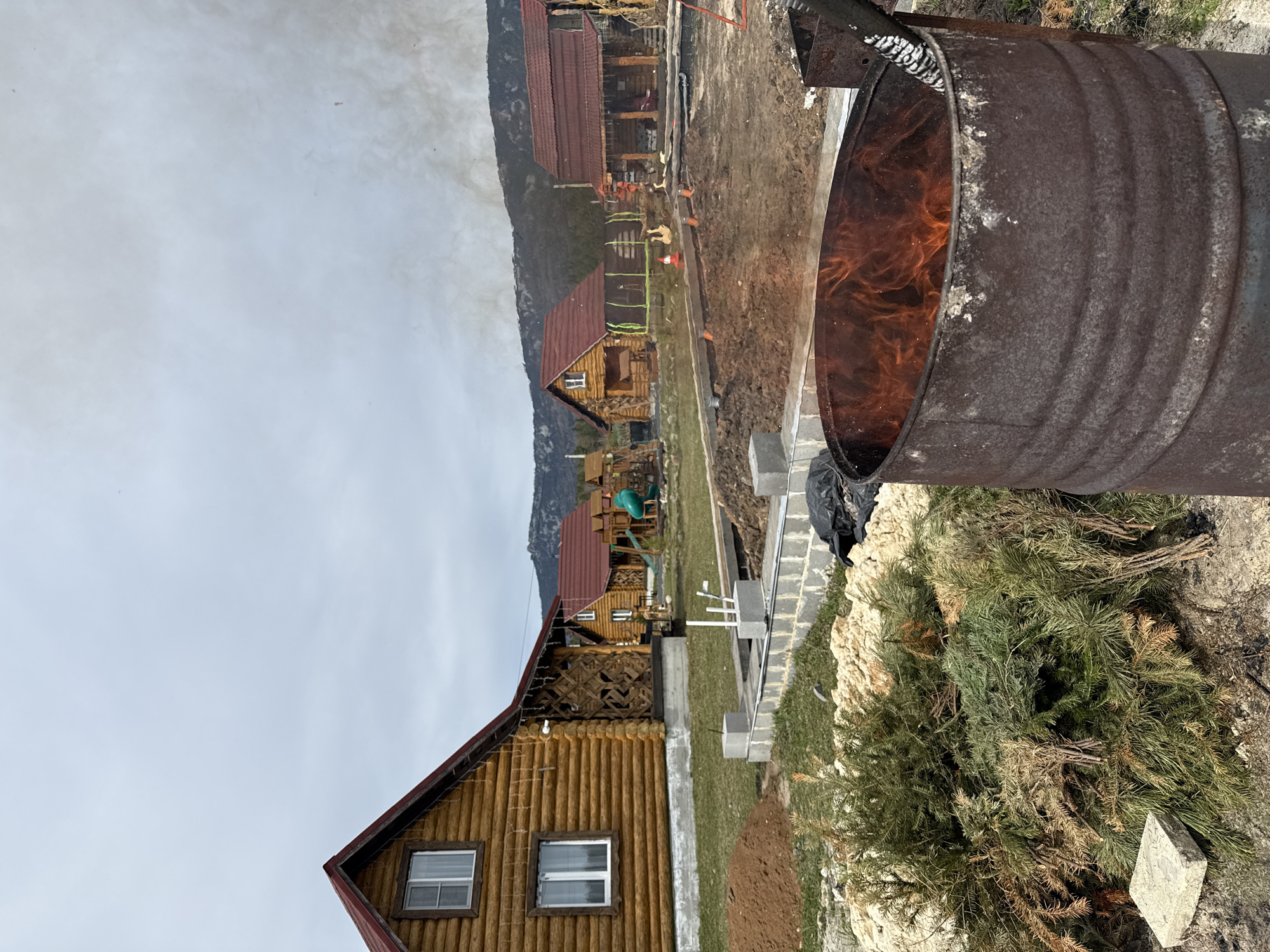 The image depicts a rural scene, likely in Tyulyuk, Russia, with log cabins visible in the background. In the foreground, a rusty metal barrel is actively burning, with flames visible inside. Piles of pine branches are lying on the ground near the barrel. Construction materials, including concrete blocks, are also present, indicating ongoing work on the property. The sky is overcast. No people are distinctly visible in the image. The overall scene suggests a construction or maintenance activity in a wooded area with rustic houses.