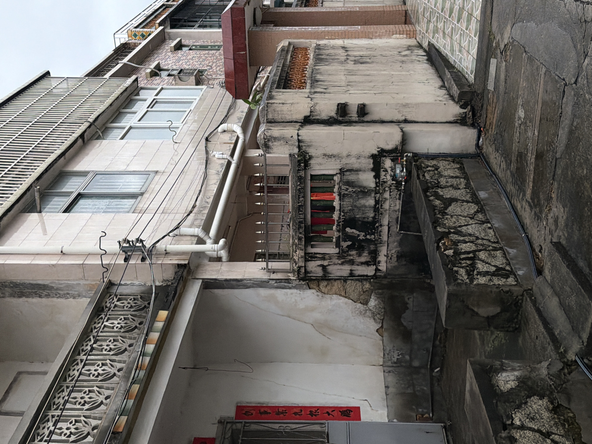 This is a medium-angle, eye-level shot of an outdoor alleyway in Longgang District, China, likely taken in the morning or late afternoon given the diffused lighting. The scene depicts the exterior of several older, multi-story buildings. The closest building on the left has a light-colored facade with visible cracks and patches of peeling paint, particularly near the base. A red banner with Chinese calligraphy is affixed to the wall to the left of a gray door.
In the mid-ground, a weathered concrete wall runs horizontally, marked with dark stains and graffiti. This wall is topped with a decorative metal railing and partially obscured by a gray metal fence. Behind this wall, elements of another building are visible, including windows with metal bars and what appears to be laundry hanging to dry in red and green colors.
Further to the right, a sidewalk with a tiled pattern, featuring green and beige squares, runs alongside a brick-clad building. A portion of a second story of this building features windows with white frames and a metal grate above them. Electrical wires are strung across the scene, connecting various points of the buildings. The ground in the foreground is a mix of cracked concrete and darker, wet-looking asphalt, suggesting recent rain. The overall impression is one of an aged urban environment with signs of wear and tear. There are no people or animals visible in the frame.