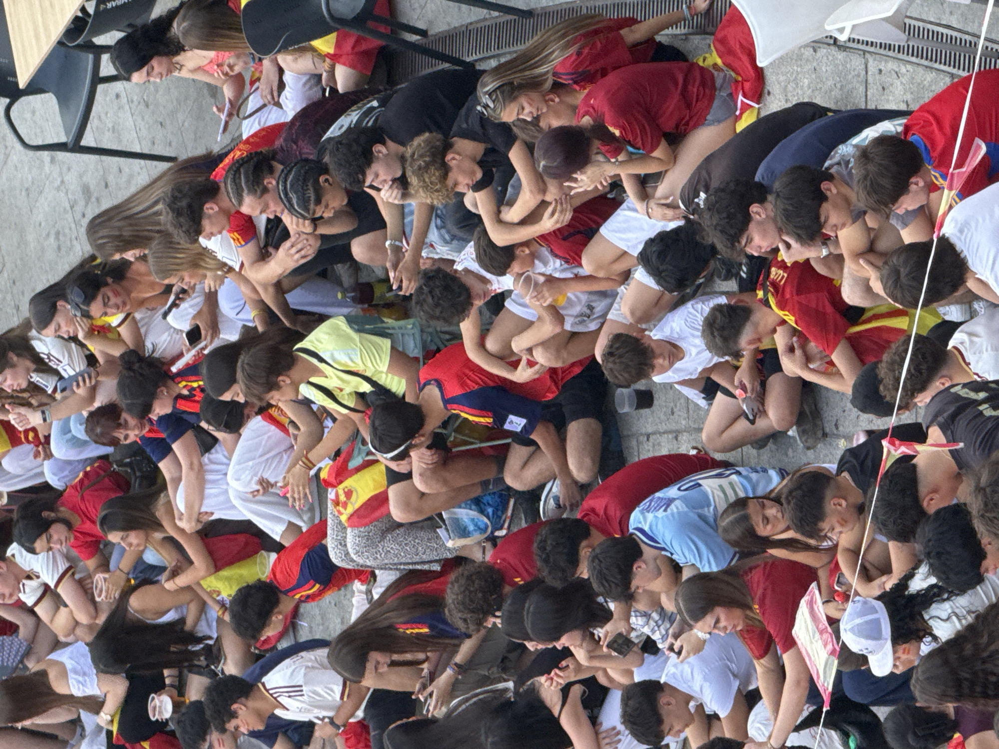This is an elevated, close-up, overhead shot of a crowd of young people, predominantly teenagers and young adults, sitting on the ground. They are gathered outdoors, possibly in a public square or plaza, as indicated by the textured paving stones and a metal grate in the lower right corner, suggesting a drain or ventilation system. Many individuals are wearing red and yellow clothing, flags, or face paint, strongly indicating a connection to a Spanish sporting event or national celebration. Several people are holding or interacting with their mobile phones. Some are holding plastic cups, suggesting they are consuming beverages. The atmosphere appears to be one of casual observation and engagement, possibly watching an event on a screen or waiting for something to happen. The clothing and hairstyles suggest a contemporary setting. While the image itself does not explicitly provide the location, the cultural indicators like the flags and clothing strongly suggest a Spanish context. A possible location in Spain, such as a public space in Las Rozas de Madrid, would be consistent with such a gathering.