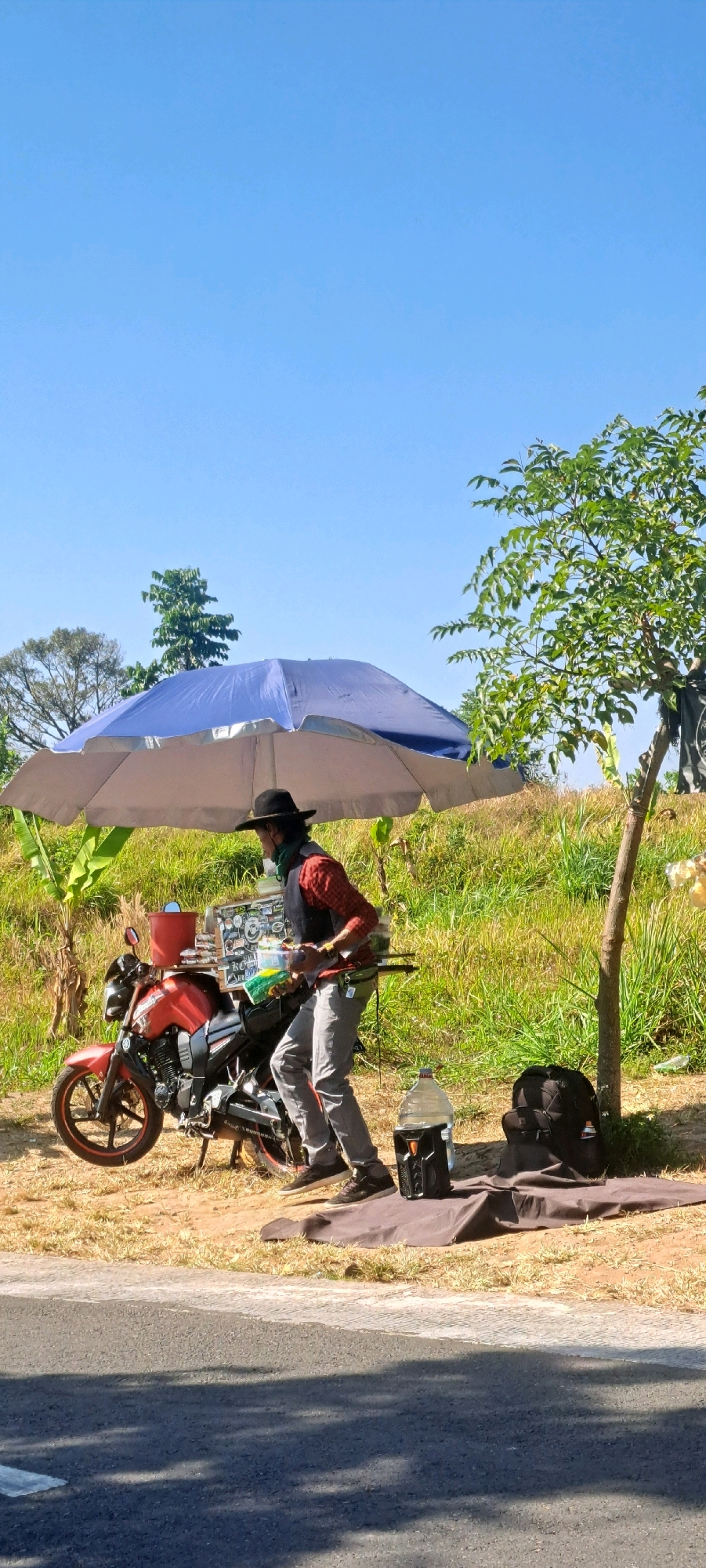 A person wearing a black hat and a red and black checkered shirt is standing next to a red motorcycle parked on a grassy area next to a road. The person appears to be preparing food or drinks, holding a green item and a white container. A large blue and silver umbrella is set up over the person and motorcycle, providing shade. On the ground near the person are a large water jug, a black backpack, and a brown mat. The environment is outdoors with green grass, trees, and a clear blue sky. The scene is situated in Sekarjoho, Indonesia.