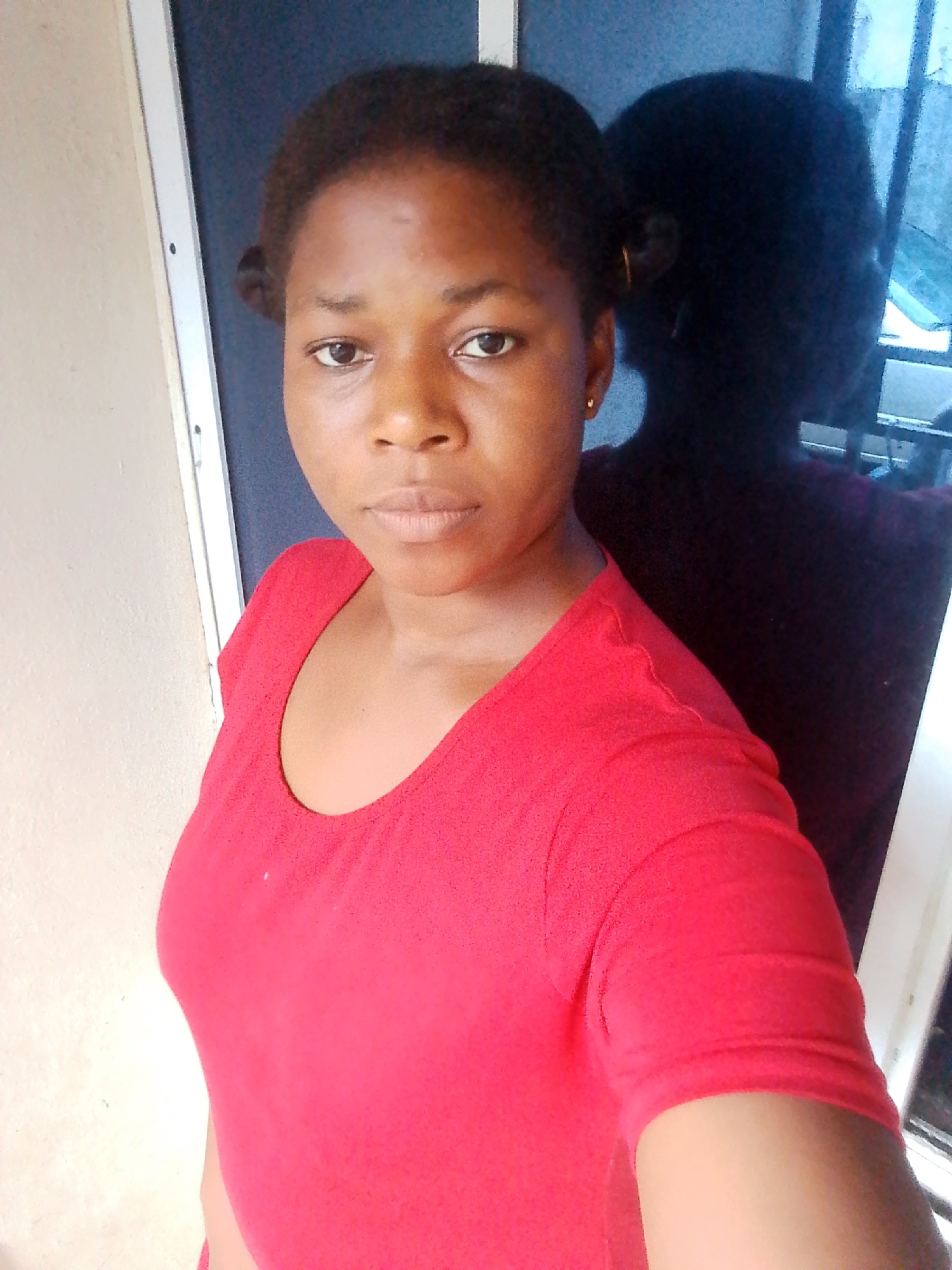 A young Black woman with dark hair pulled into two buns is taking a selfie. She is wearing a bright red t-shirt with a V-neckline. Her skin tone is medium to dark, and she has dark eyes. A faint shadow of another person is visible behind her, reflected in a window. The background includes a light-colored wall and a dark doorway. The image appears to be taken indoors, possibly in a home, and the lighting suggests daytime. The woman has a neutral expression. There is no visible text in the image.