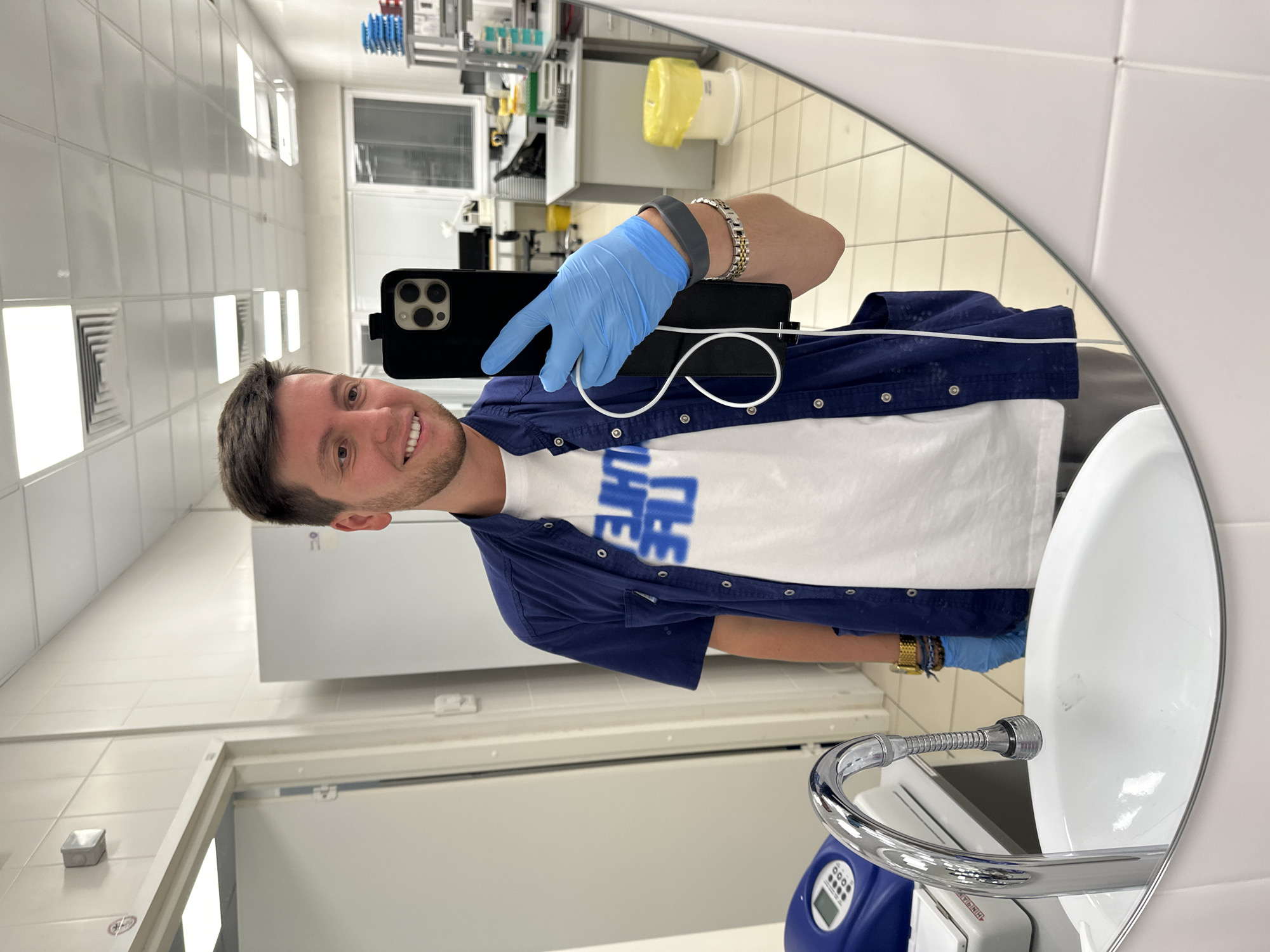 A man with short brown hair and a light beard, wearing a dark blue open short-sleeved shirt over a white t-shirt with blue lettering and light grey pants, stands in a laboratory environment and takes a mirror selfie. He is wearing blue nitrile gloves and a grey fitness tracker on his left wrist, along with a watch and a bracelet on his right wrist. He holds a black smartphone in his left hand, with the camera facing the mirror. The laboratory features white tiled walls, fluorescent ceiling lights, lab benches, equipment, and a sink with a chrome faucet. The man is smiling and looking at his reflection. The scene is set in a laboratory in Moscow, Russia.