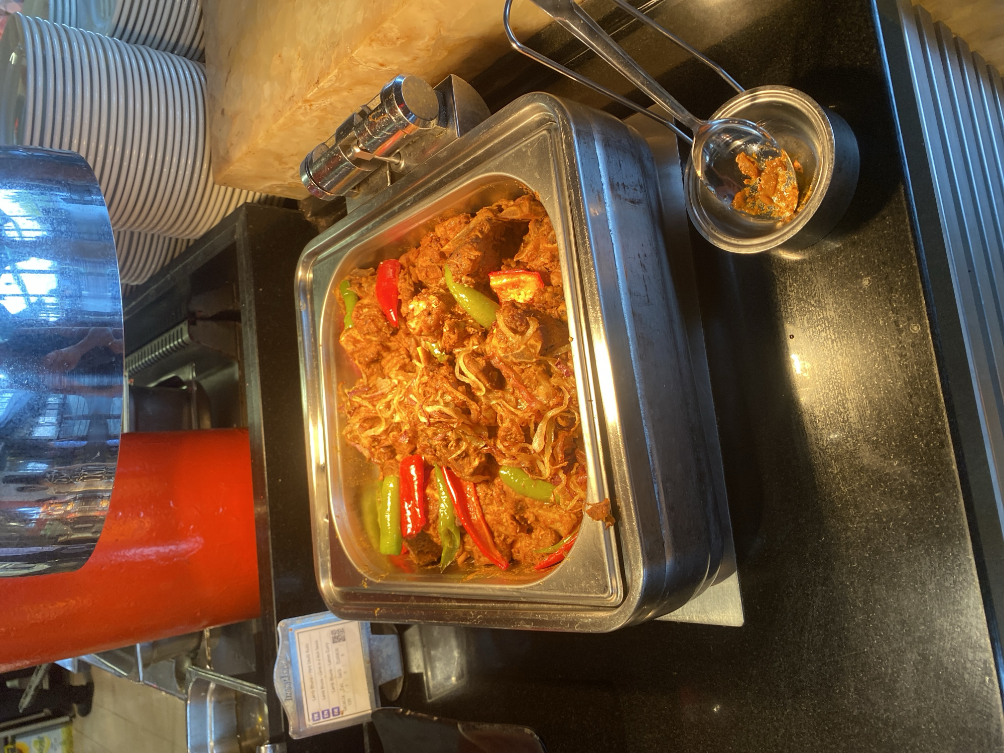 The image shows a close-up of a buffet serving dish filled with a red-colored curry, likely lamb, garnished with red and green chili peppers and topped with fried onions. The serving dish is made of stainless steel and is placed on a black granite countertop. To the right of the serving dish, there is a small metal container with a ladle holding some of the curry. In the background, there are stacks of white plates and a red buffet dispenser. A small information card is visible to the left of the serving dish, indicating that this is likely a food establishment. The scene is brightly lit, suggesting an indoor setting, possibly a restaurant or hotel buffet. There are no people visible in the image. The image does not provide any information about the city of Aksu, Turkey.