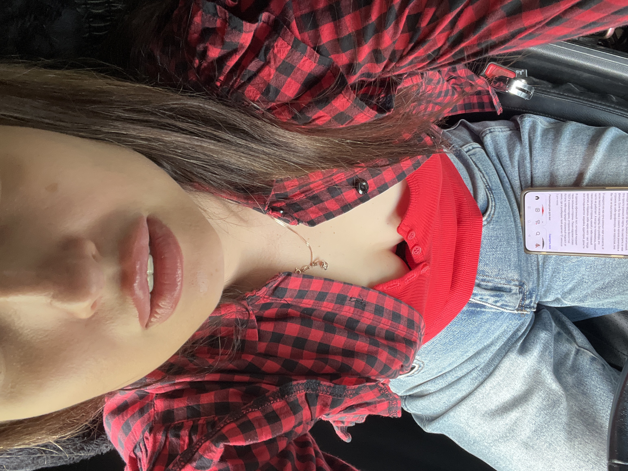 The image captures a close-up, high-angle view of an individual, likely female, from the lower face down to the lap, situated within the interior of a vehicle. The person has light skin, plump lips that are slightly parted, and long, dark brown hair falling over the shoulders. They are dressed in a red and black buffalo plaid long-sleeved button-up shirt worn open over a red ribbed knit top with a button placket, partially exposing the cleavage. A delicate gold-colored necklace with a small charm is visible around the neck. Light-wash blue jeans cover the lower body. A smartphone with an active screen displaying text in Cyrillic characters (e.g., "Смотреть все", "Рекомендации") is held in one hand and rests on the jeans. A silver car seatbelt buckle mechanism with a red release button is visible on the right side, set against a dark, likely leather, car interior. The background is predominantly dark, indicating the confines of a vehicle. The location is Unknown, Unknown.