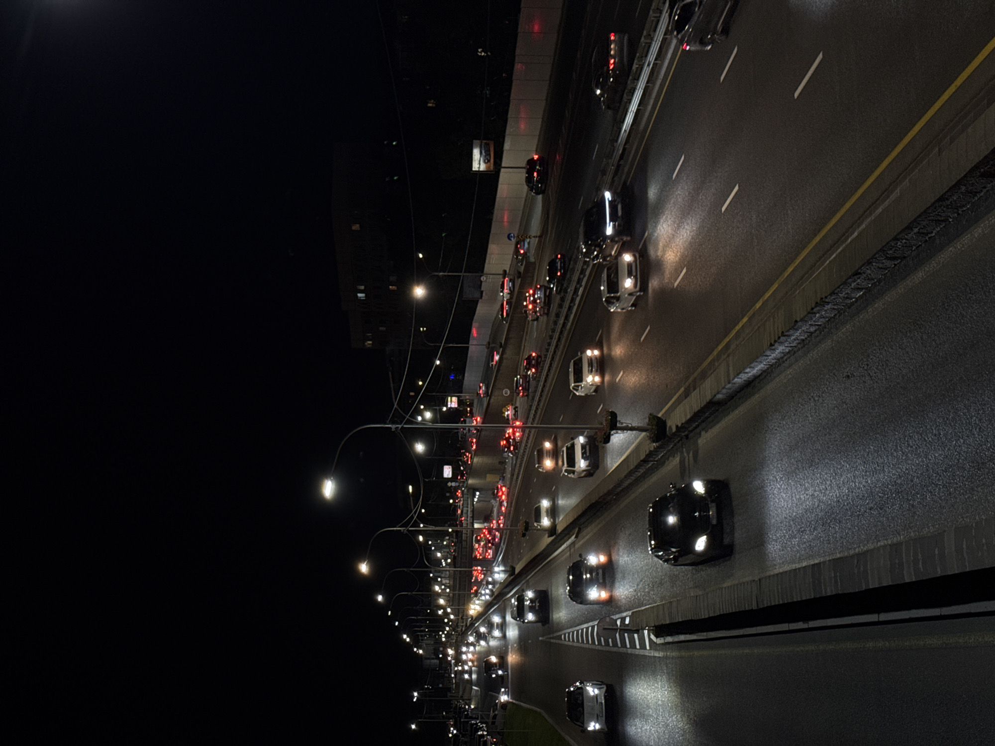 A nighttime, elevated view captures a multi-lane highway in Almaty, Kazakhstan, illuminated by streetlights and the headlights and taillights of numerous vehicles. The highway has multiple lanes of traffic moving in both directions, separated by concrete barriers and yellow and white lane markings. Cars are the primary subjects, with headlights and taillights creating streaks of light on the wet asphalt. In the distance, a multi-story building with illuminated windows is visible against the dark sky. The overall scene depicts a busy urban transportation artery at night.