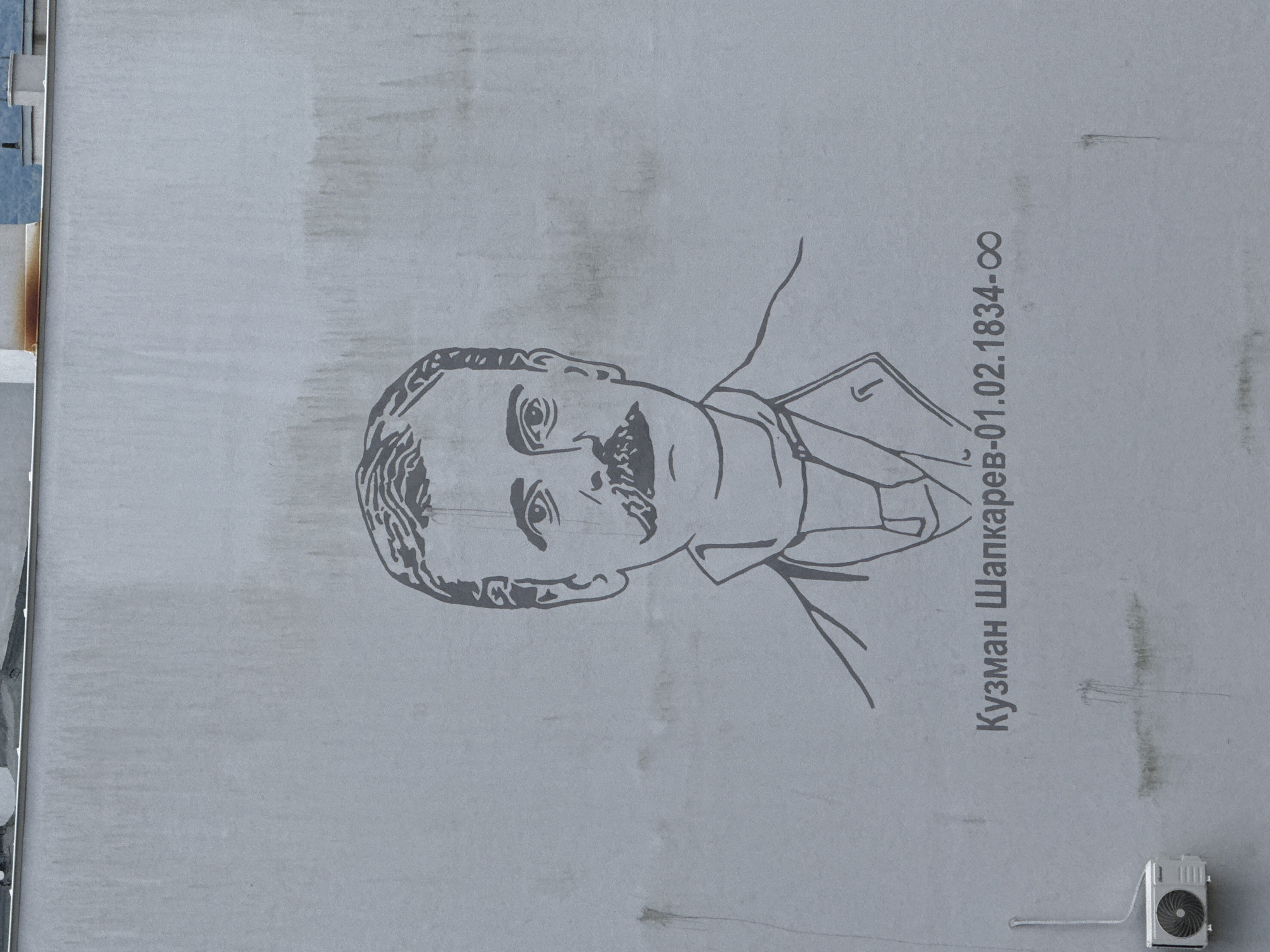 A light gray building wall in Blagoevgrad, Bulgaria, features a minimalist black line drawing of a man from the chest up, centered on the wall. The man has a prominent mustache and a stern expression, facing forward. He is depicted wearing a collared shirt and what appears to be a jacket or coat. Below the drawing, in dark gray Cyrillic script, is the text "Кузман Шапкарев-01.02.1834-∞". The wall surface exhibits vertical streaks and patches of discoloration consistent with dirt and water runoff. In the lower-left corner, a white outdoor air conditioning condenser unit is mounted, with a thin white conduit extending upwards from it. The top edge of the image reveals a dark horizontal line, likely a rooftop or parapet, and faint, blurred elements in the distant background.