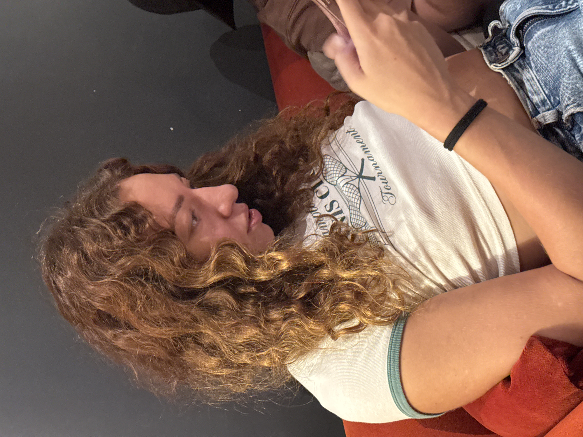 A young woman with curly brown hair sits on a red couch, looking down at her phone. She is wearing a white crop top with a green trim and blue denim shorts. A black hair tie is wrapped around her wrist. The background is a dark, solid wall. The image does not provide any information about the location in Hanoi, Vietnam.