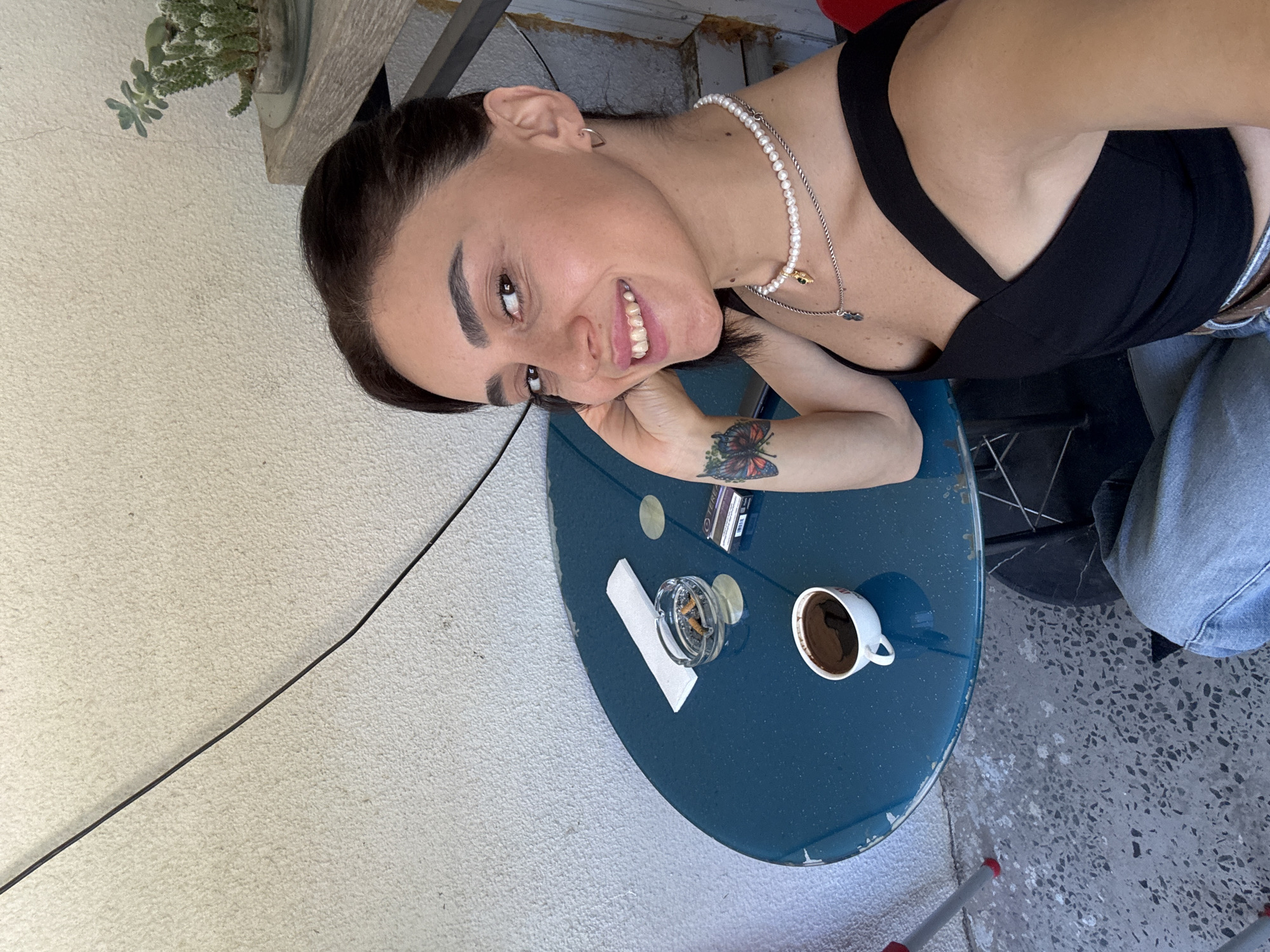 A woman sits at a round, blue table outdoors. She is facing the camera, smiling, and looking slightly to her left. She has dark hair pulled back, and is wearing a black top with criss-cross straps and a pearl necklace. On her left wrist, she has a tattoo of a butterfly. On the table in front of her is a white coffee cup, an ashtray with cigarette butts, a pack of cigarettes, and a white napkin. The table is set against a textured, off-white wall. Behind the woman, a small succulent plant sits on a shelf. In the foreground, the woman's legs, clad in blue jeans, are visible. The image appears to have been taken in Tbilisi, Georgia.